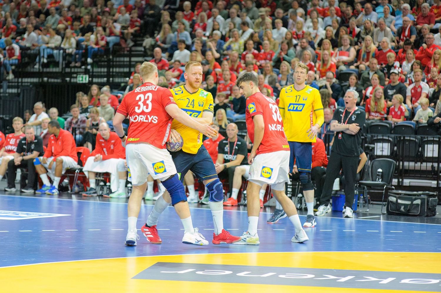 Professional handball match action shows players in red and yellow jerseys competing on a blue indoor court.
