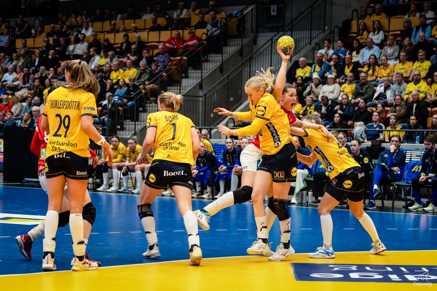 Female handball players in yellow uniforms jump to block a shot during an intense indoor match.