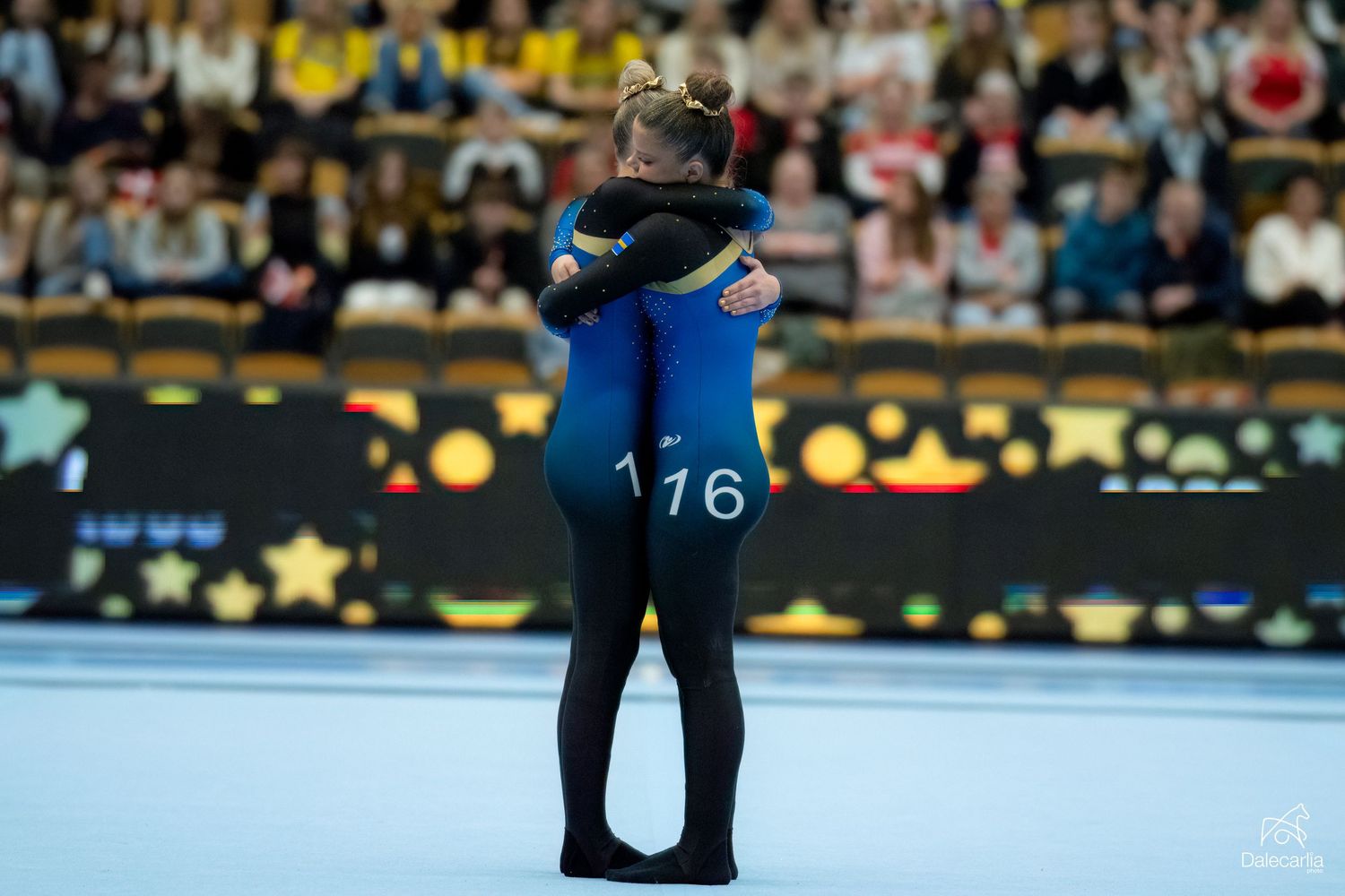 Two figure skaters embrace on the ice during a competitive performance while spectators watch from the stands.