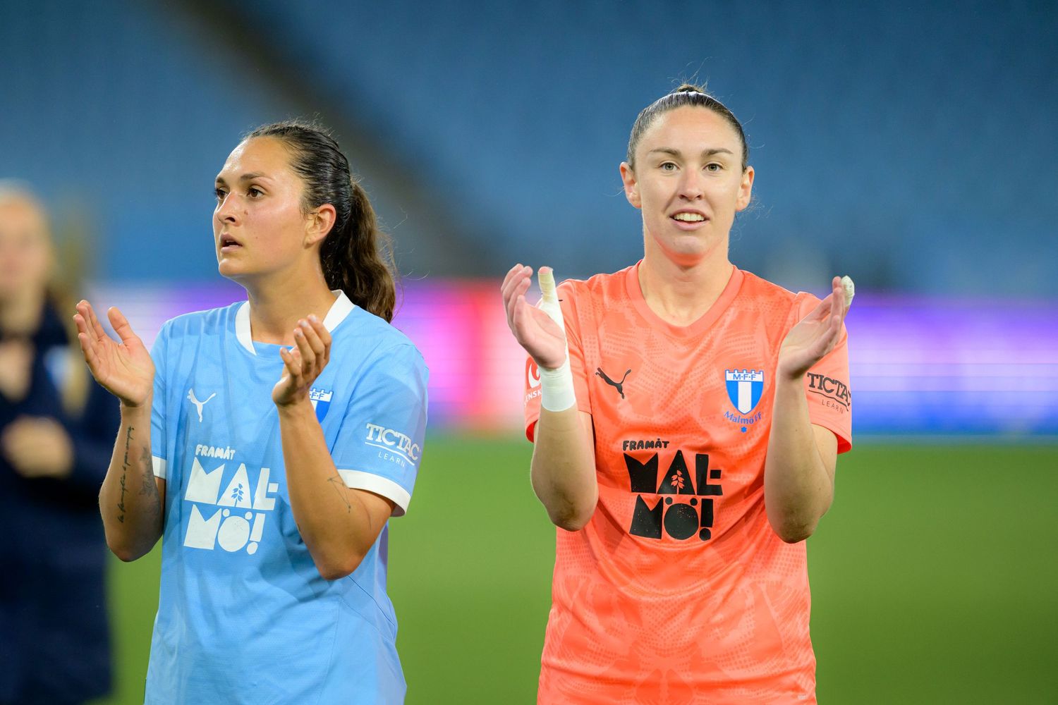 Two soccer players in light blue and coral jerseys applaud while standing on a field during a match.