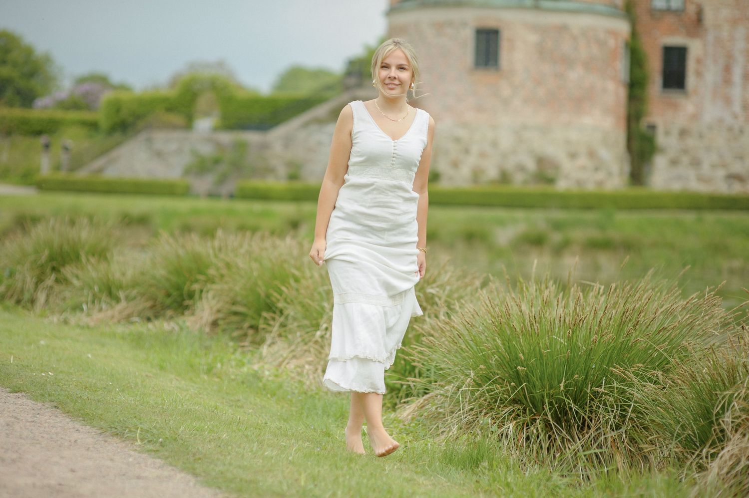 A person in a flowing white dress walks barefoot along a path near an old stone building on a summer day.