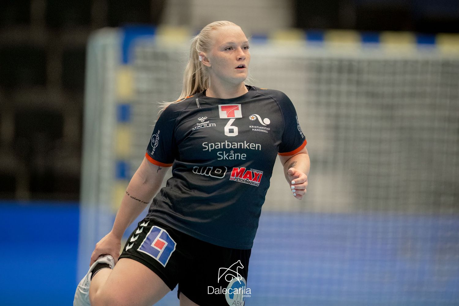 A handball athlete in a dark jersey runs on an indoor court with blue flooring and goal visible in background.