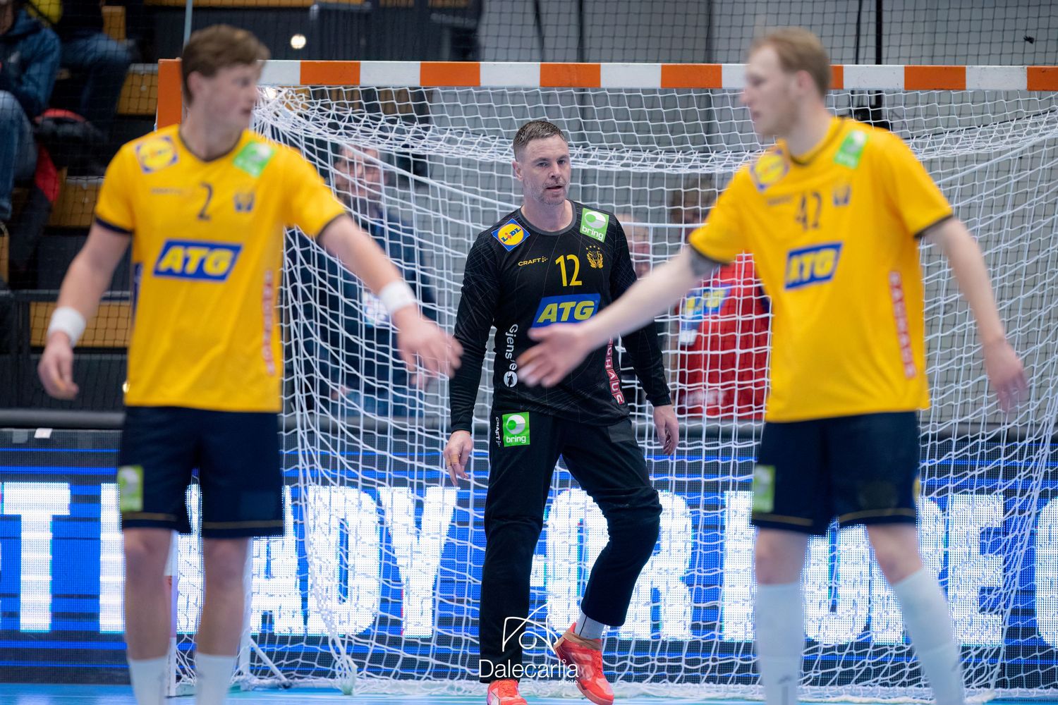 Handball match scene with two players in yellow jerseys and a goalkeeper in black uniform standing near the goal net.