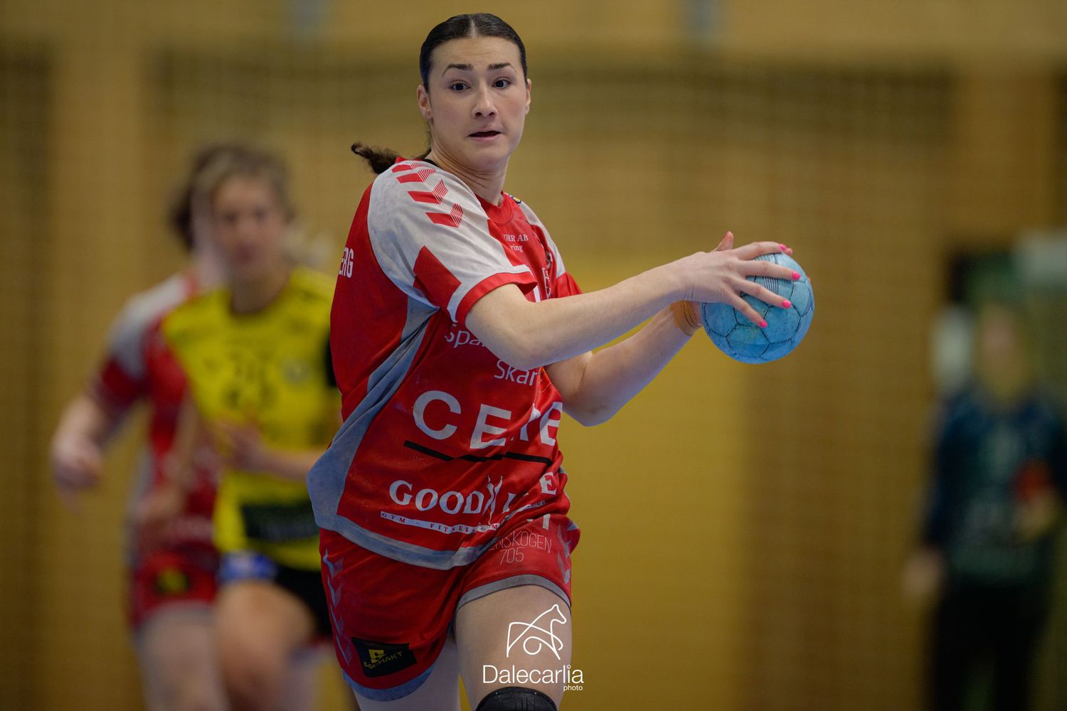 A handball player in red and white jersey extends arm while holding a blue ball during indoor match.