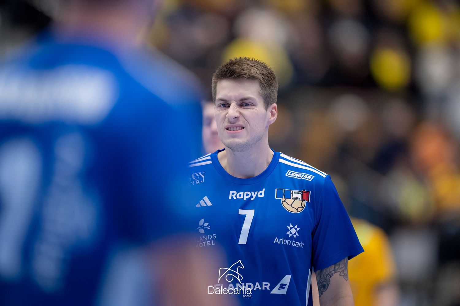 Handball player wearing a blue Flensburg team jersey with number 7 watches the action during a match.