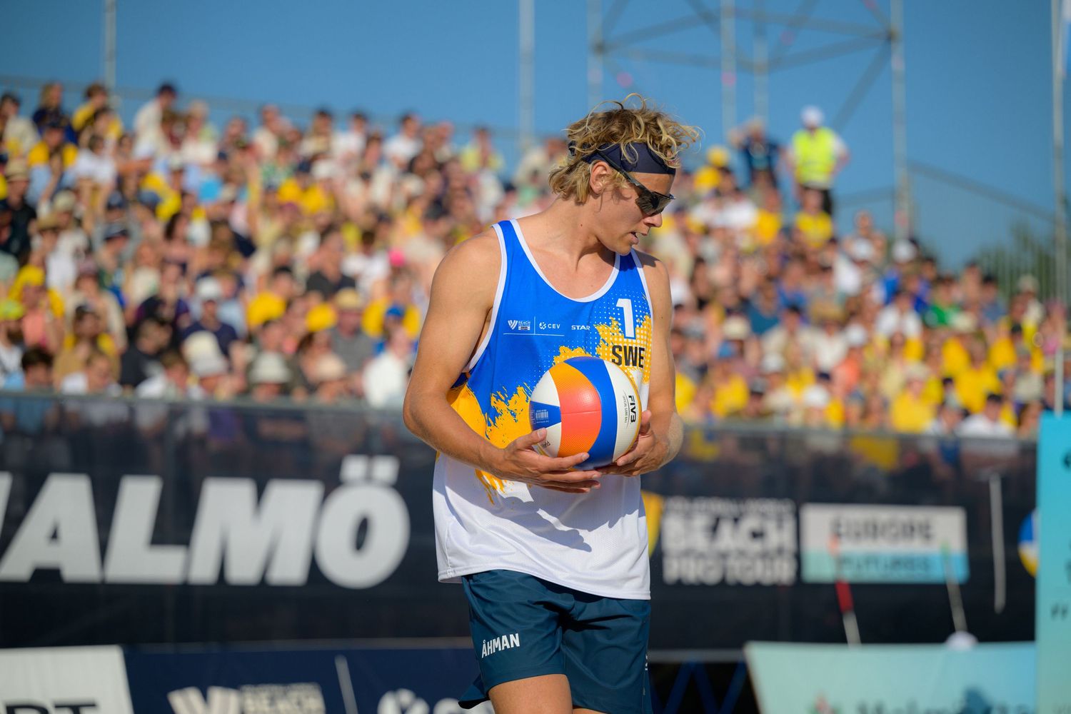 An athlete in blue and white jersey competes during a beach volleyball match in front of a crowded stadium.