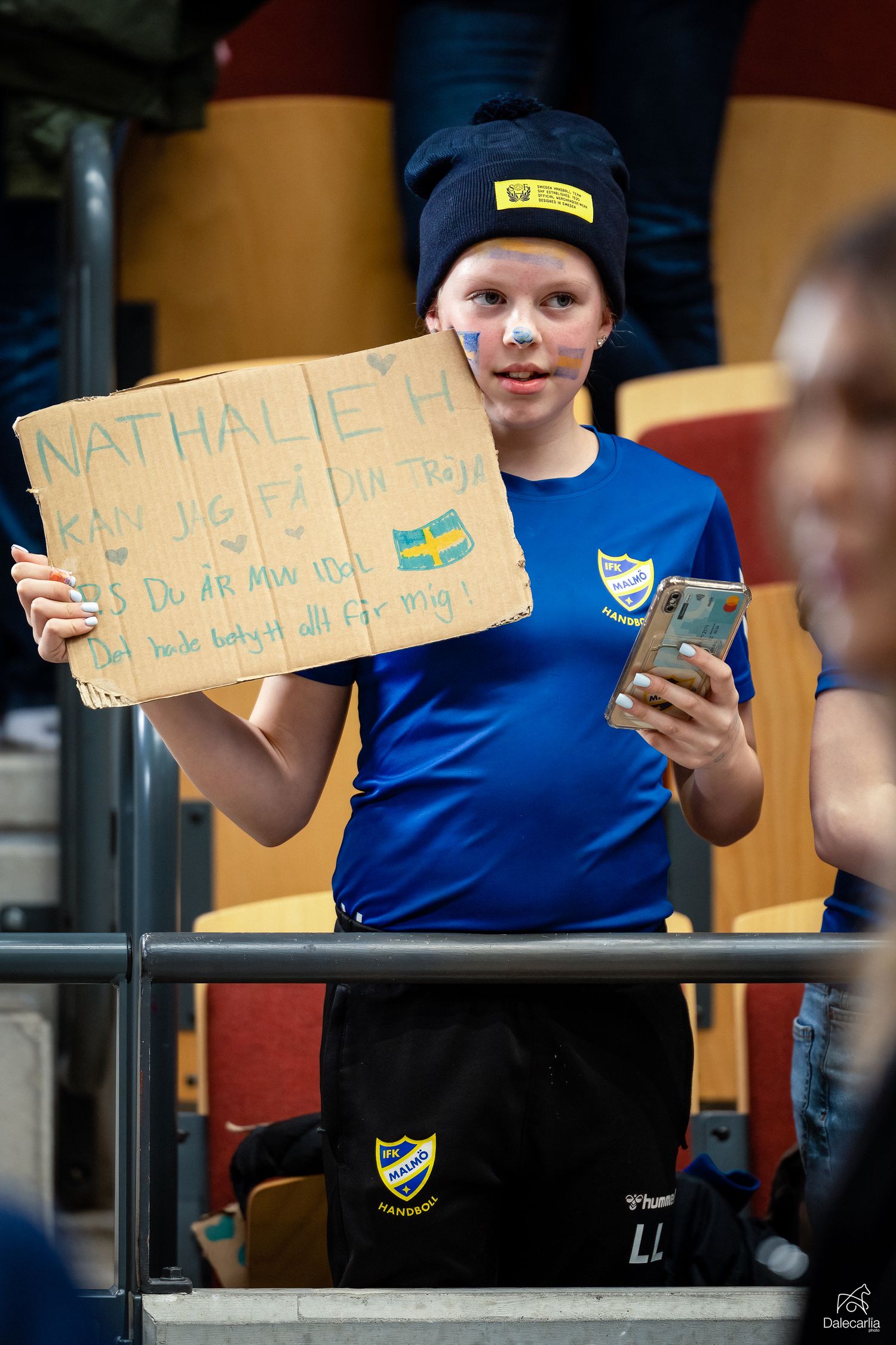 A young sports fan in blue uniform holds up a homemade sign and money at what appears to be a sporting event in Sweden.
