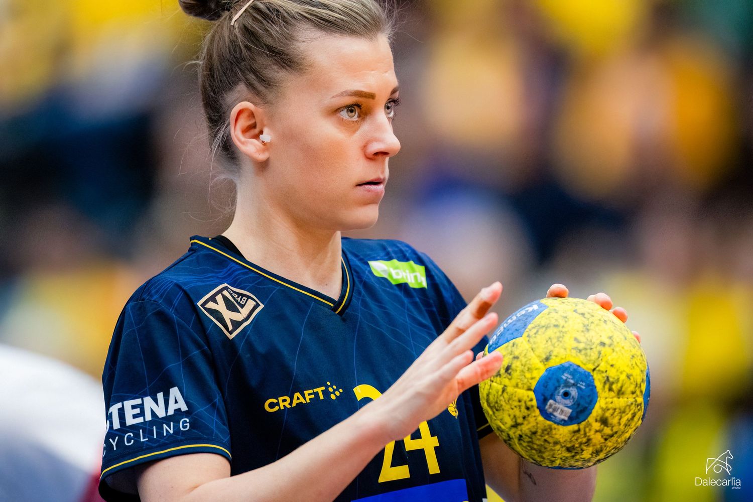Handball player in dark blue jersey holds yellow ball during competitive match with yellow background.