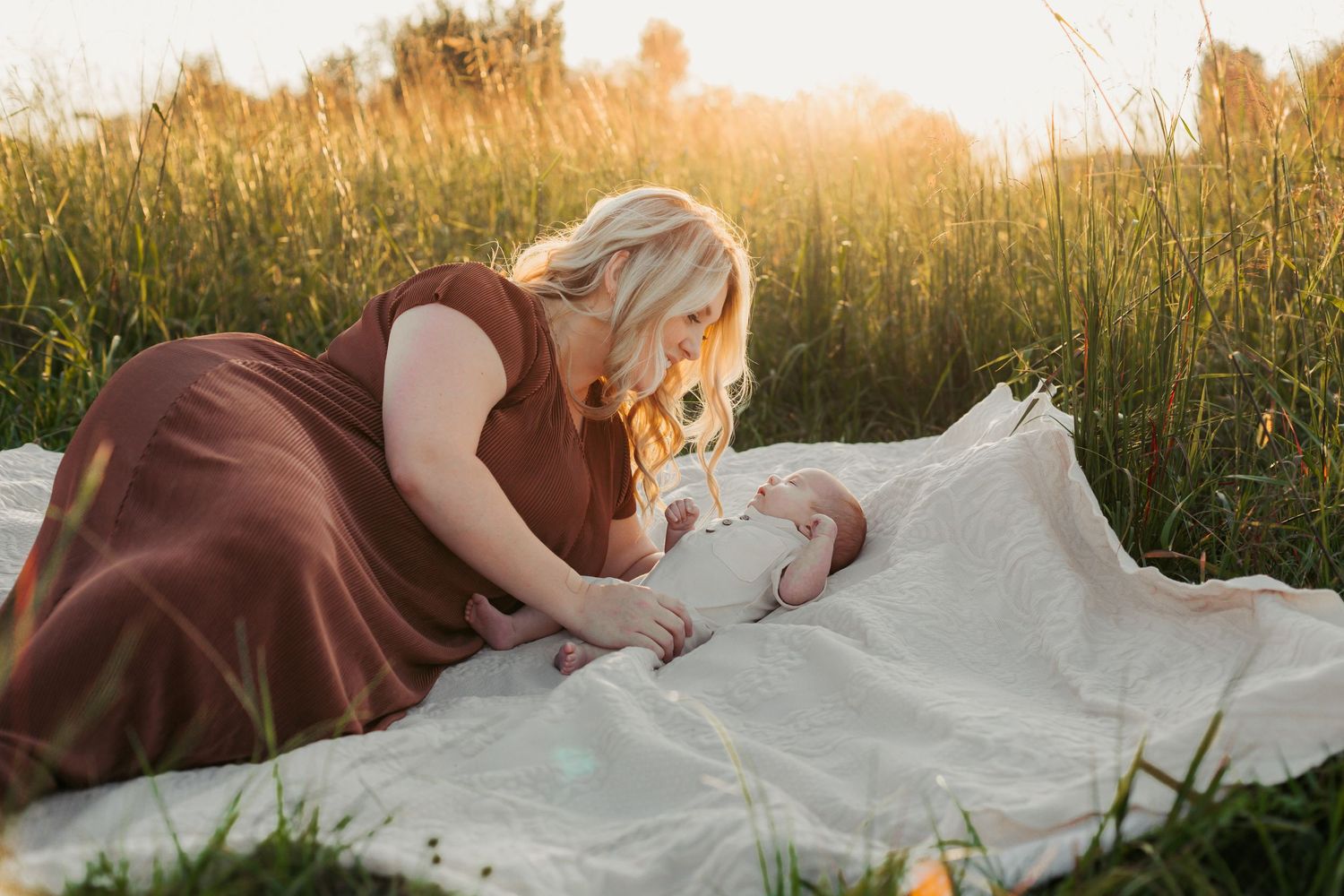 Mother and baby share tender moment on blanket in sunlit meadow.