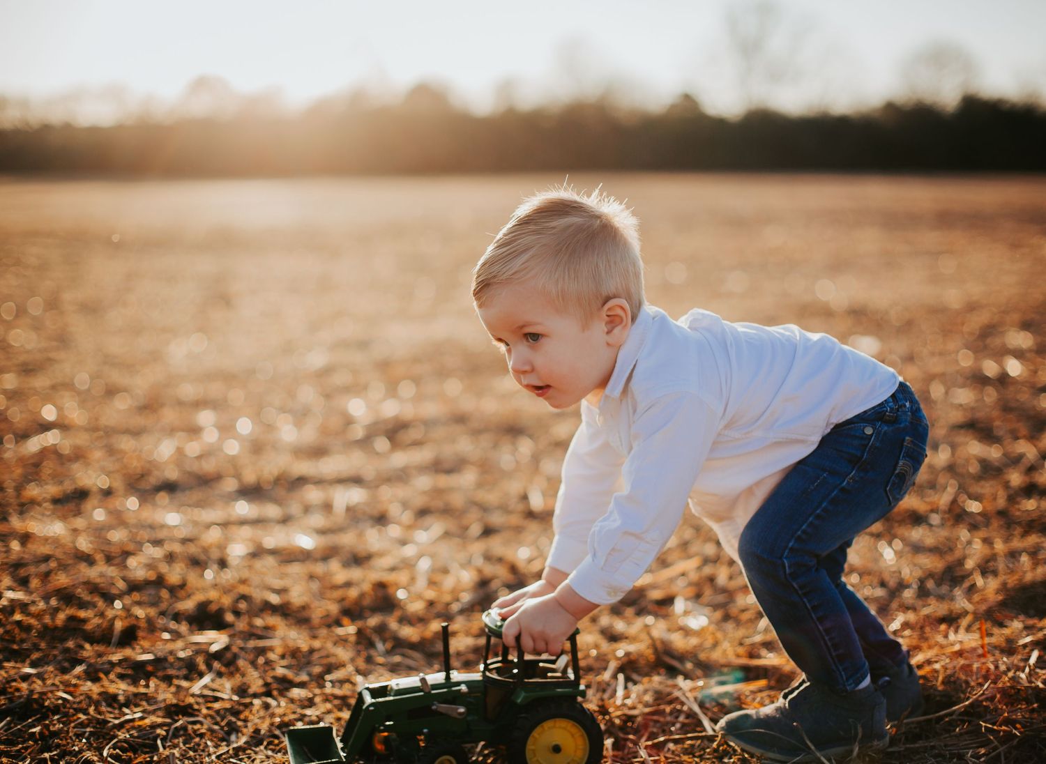 Child bends down to play with green toy tractor in autumn field at sunset.