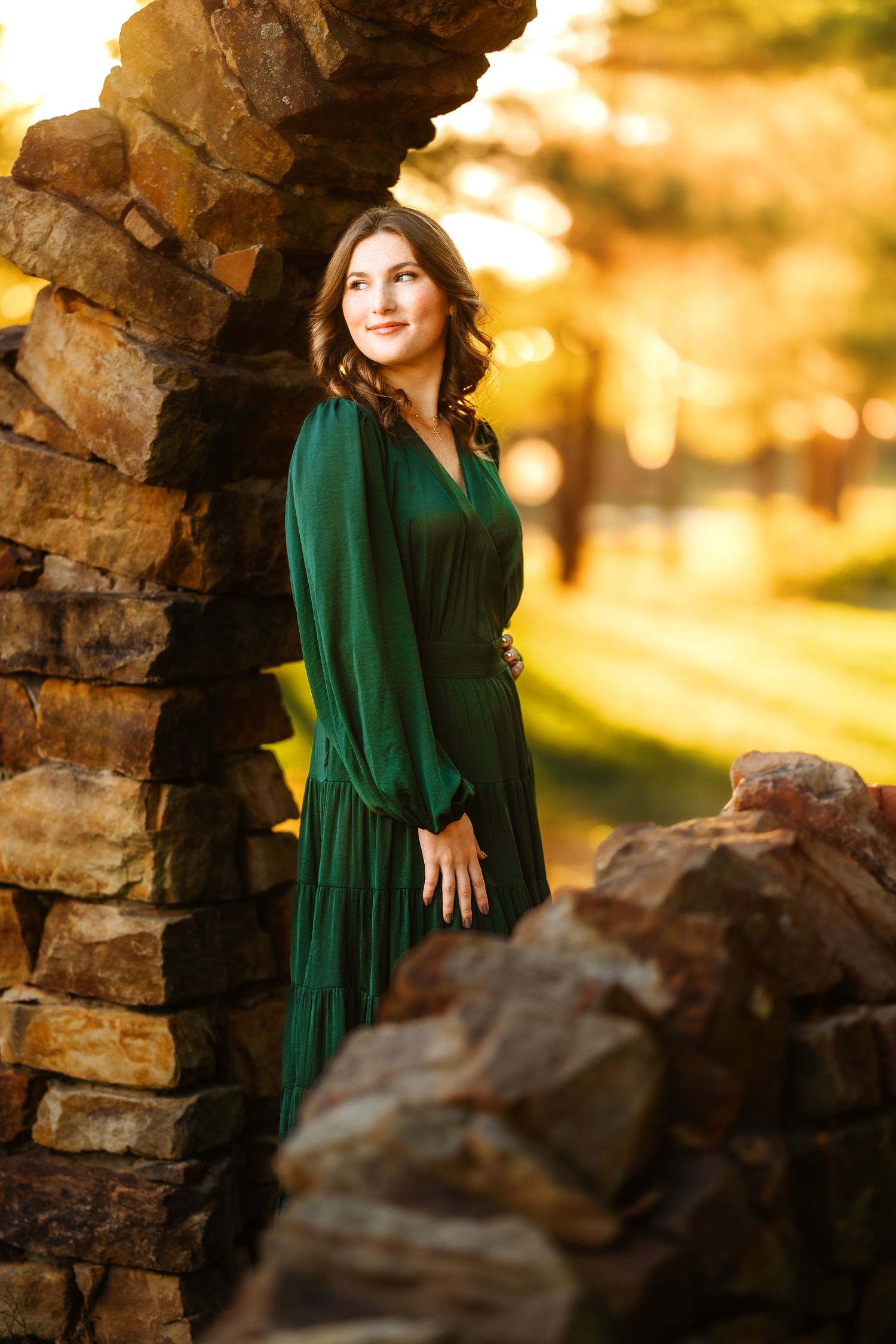 Portrait of person in emerald green dress leaning against stone arch during golden sunset in autumn.