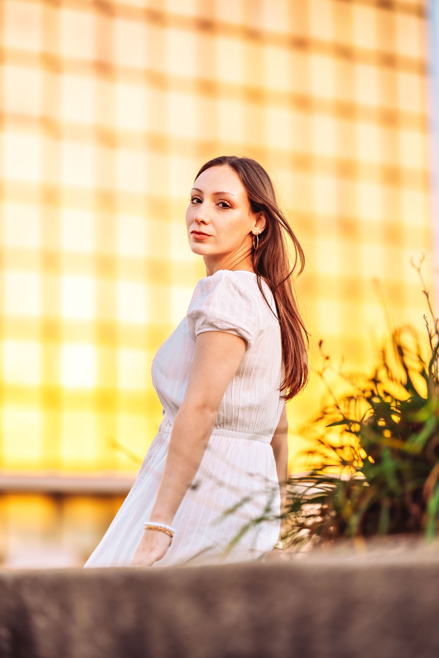 Portrait of person in white dress posing outdoors at sunset with yellow illuminated building in background.