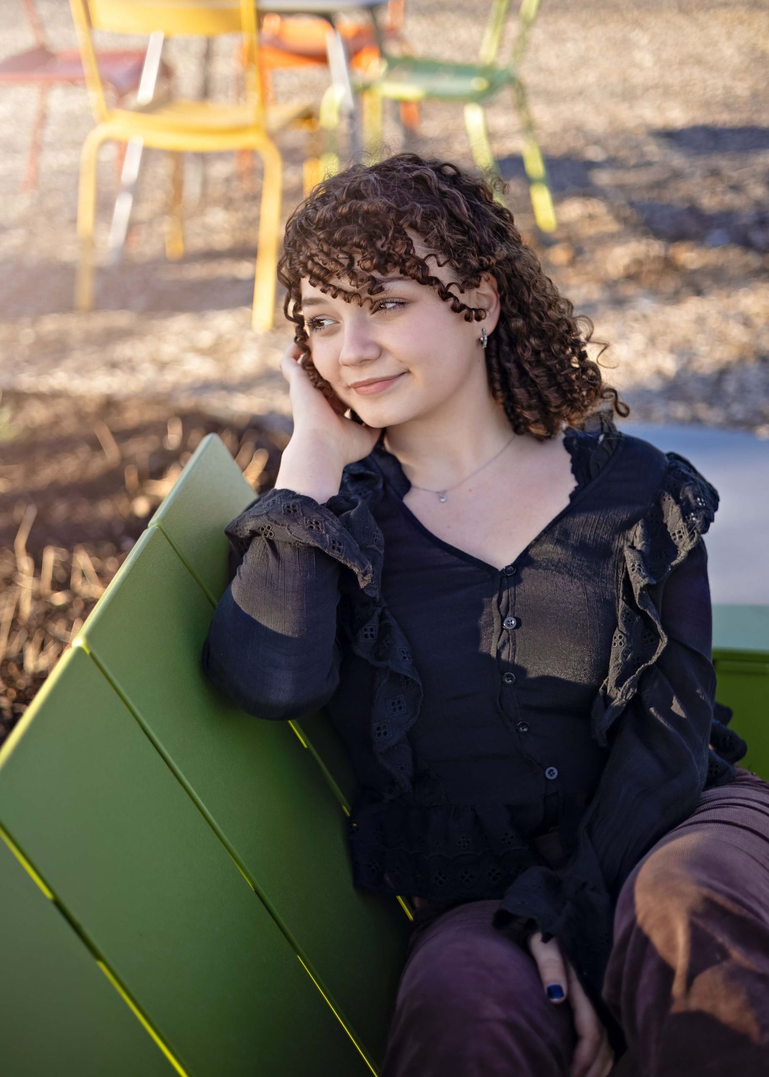 A young person with curly dark hair wearing a black ruffled top sits on a green bench in outdoor lighting.