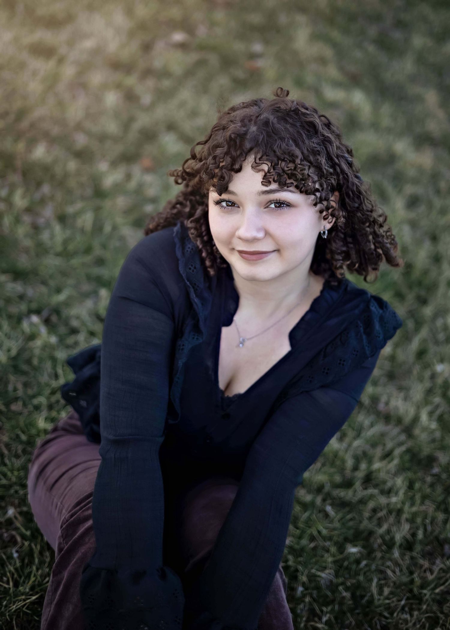 Smiling individual with curly dark hair in a black top sitting on grass during an outdoor portrait session.