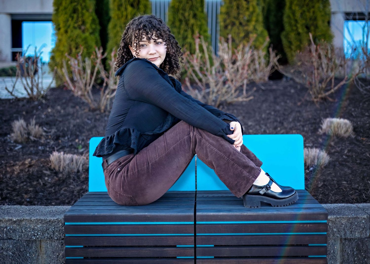 Person wearing black shirt and brown pants sits on bright blue bench outdoors with winter shrubbery in background.