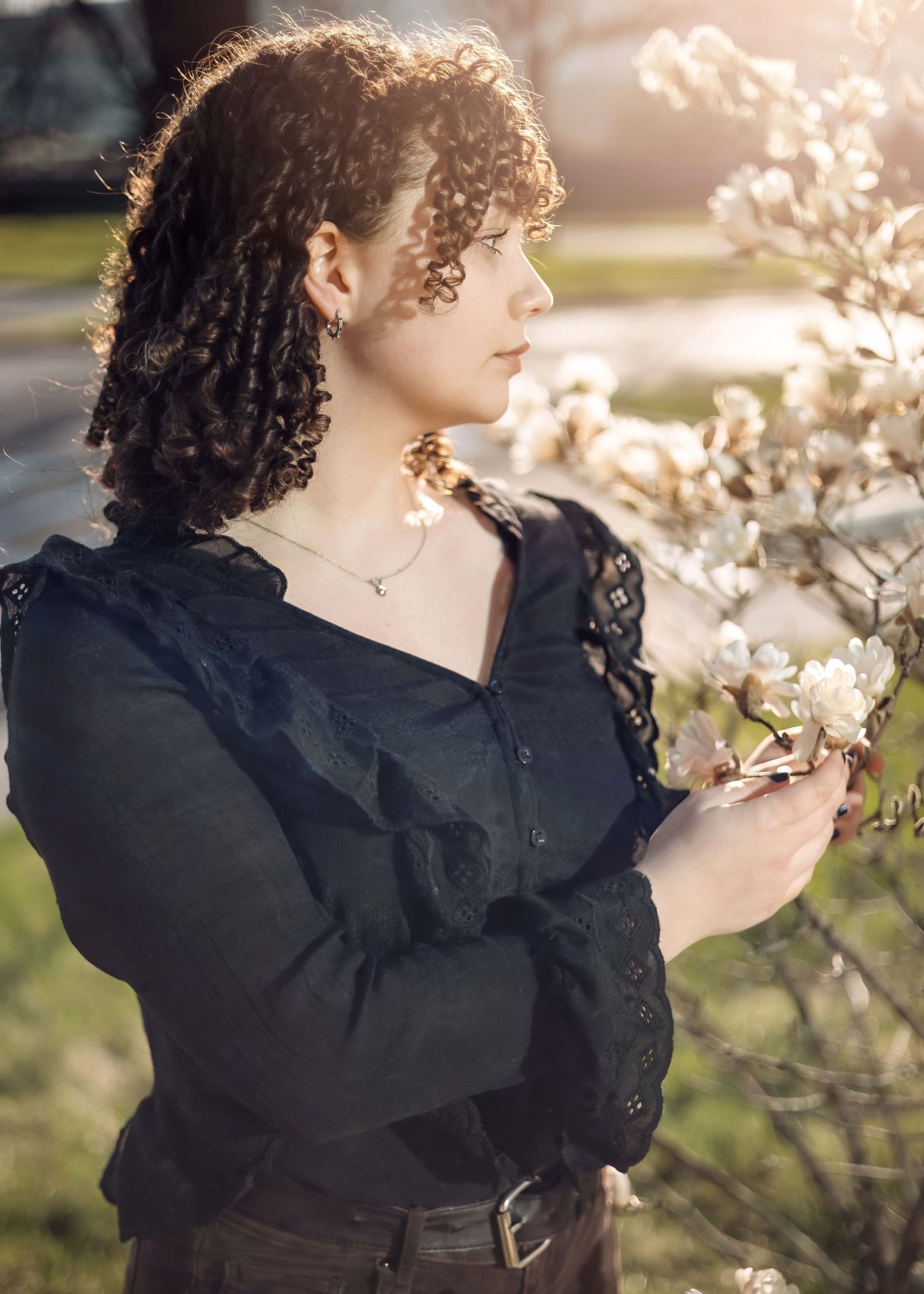Person with curly dark hair wearing a black top admires blooming white flowers in soft sunlight.