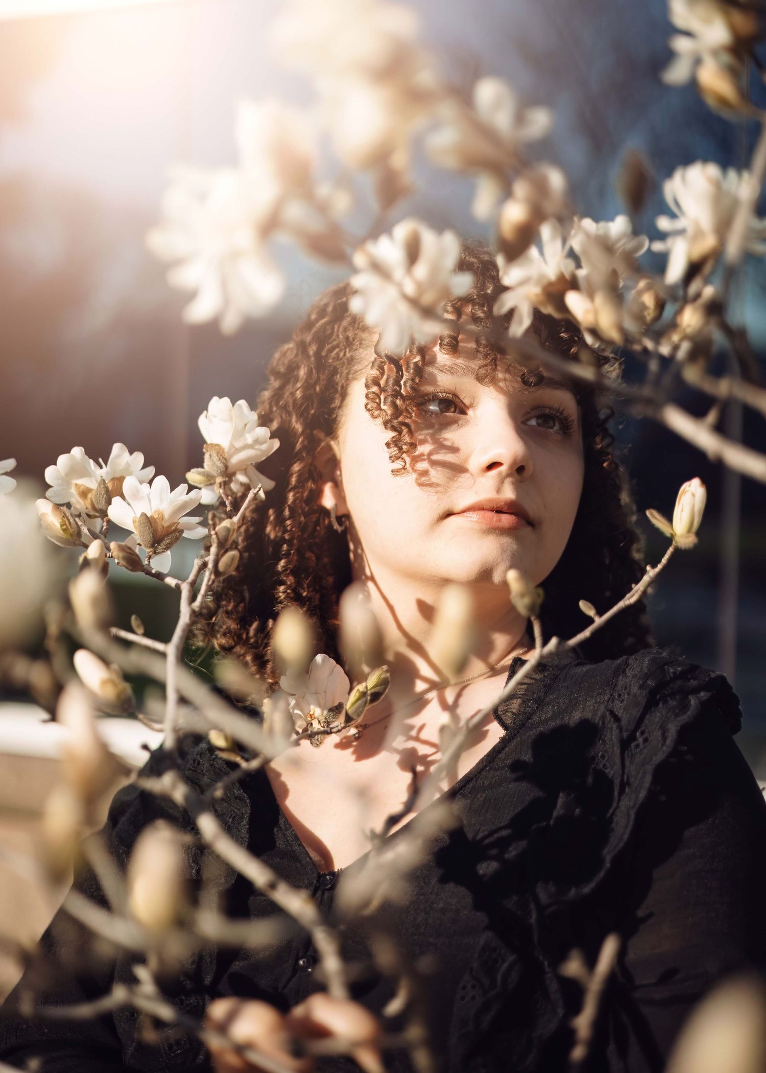 A romantic spring portrait with white flower blossoms framing a figure in black against soft sunlit bokeh.