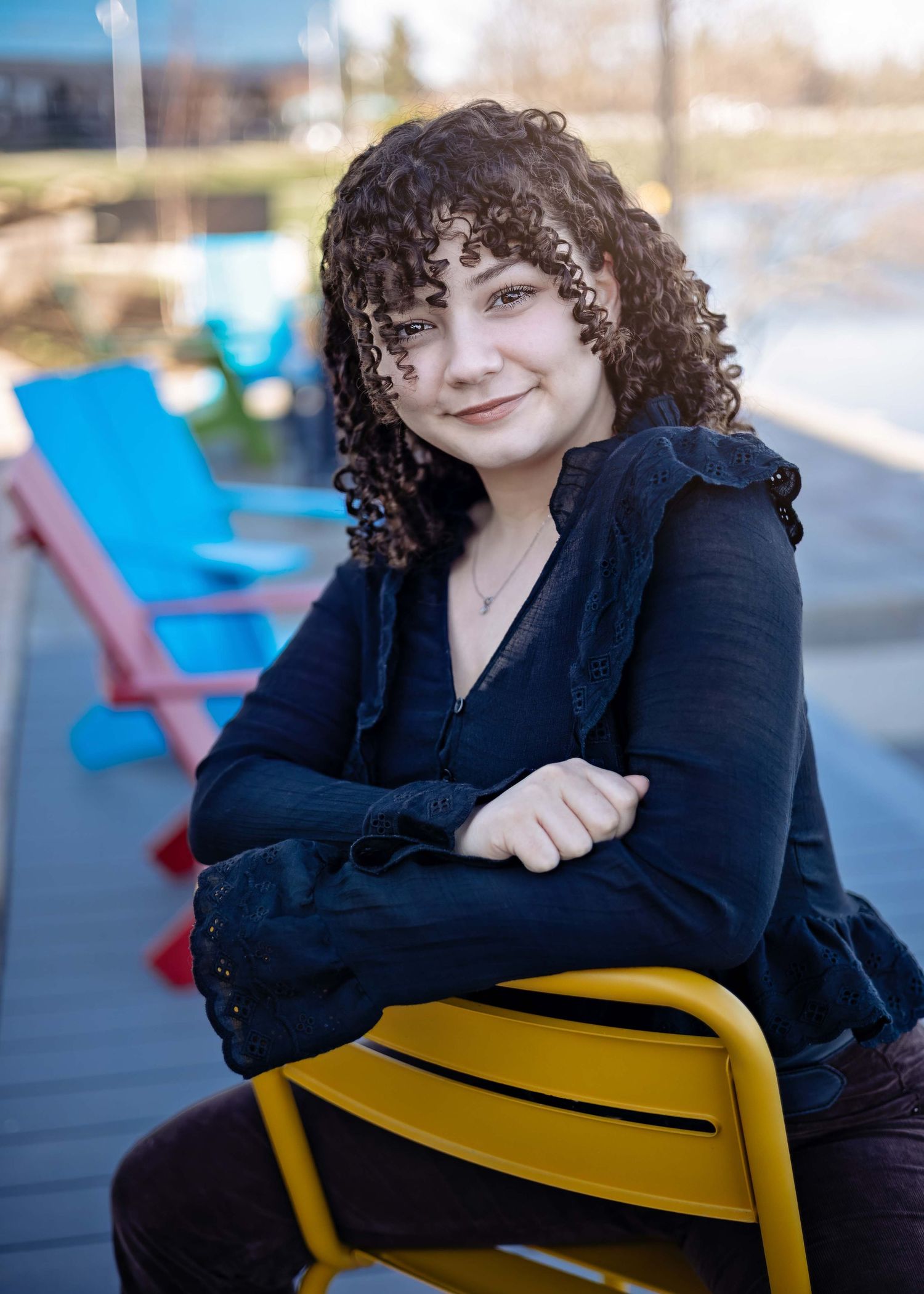 A person sits on a yellow chair outdoors, wearing a black shirt and smiling warmly with curly hair.
