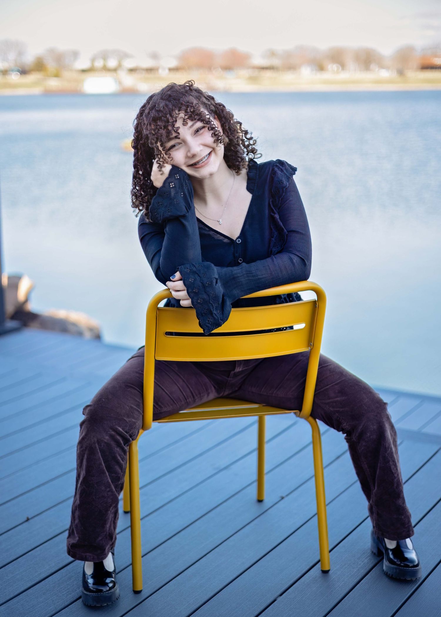 A person in a black top sits casually on a yellow metal chair on a deck overlooking a waterfront view.