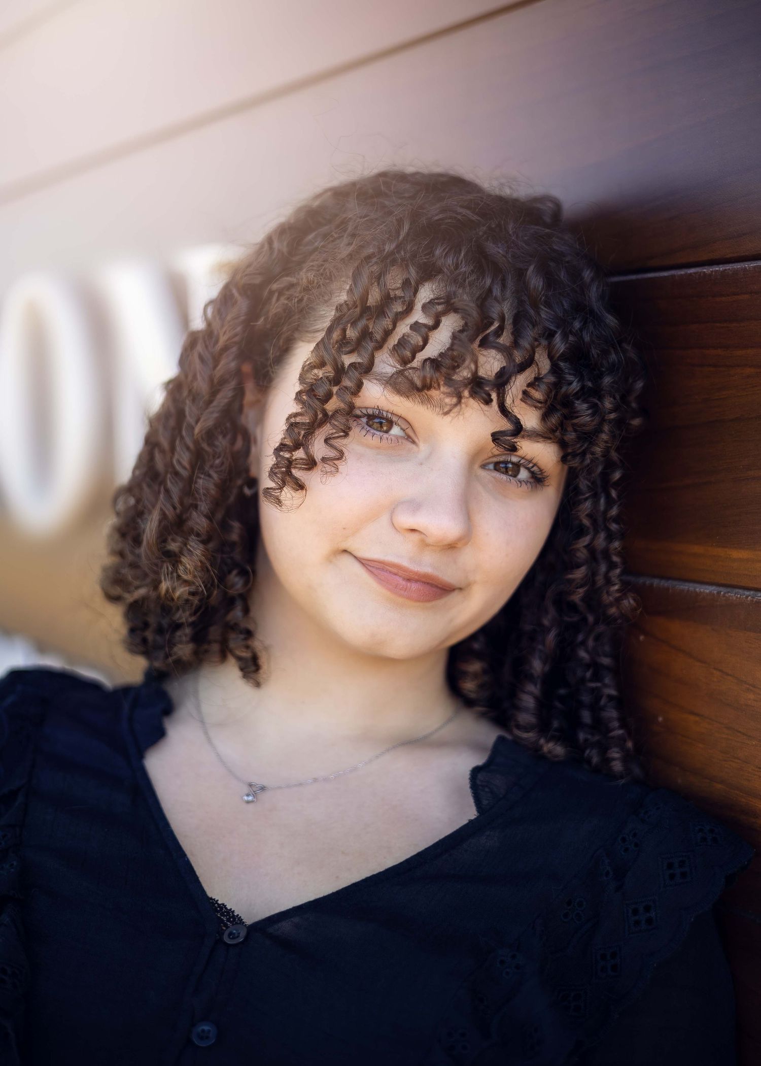 Portrait of someone with natural curly dark hair and bangs wearing a black top against a wooden wall.