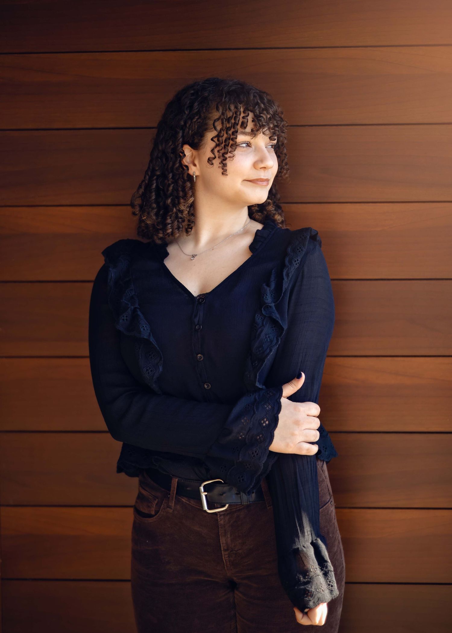 Someone with dark curly hair and bangs wearing a black ruffled blouse and brown pants poses against a wooden wall.