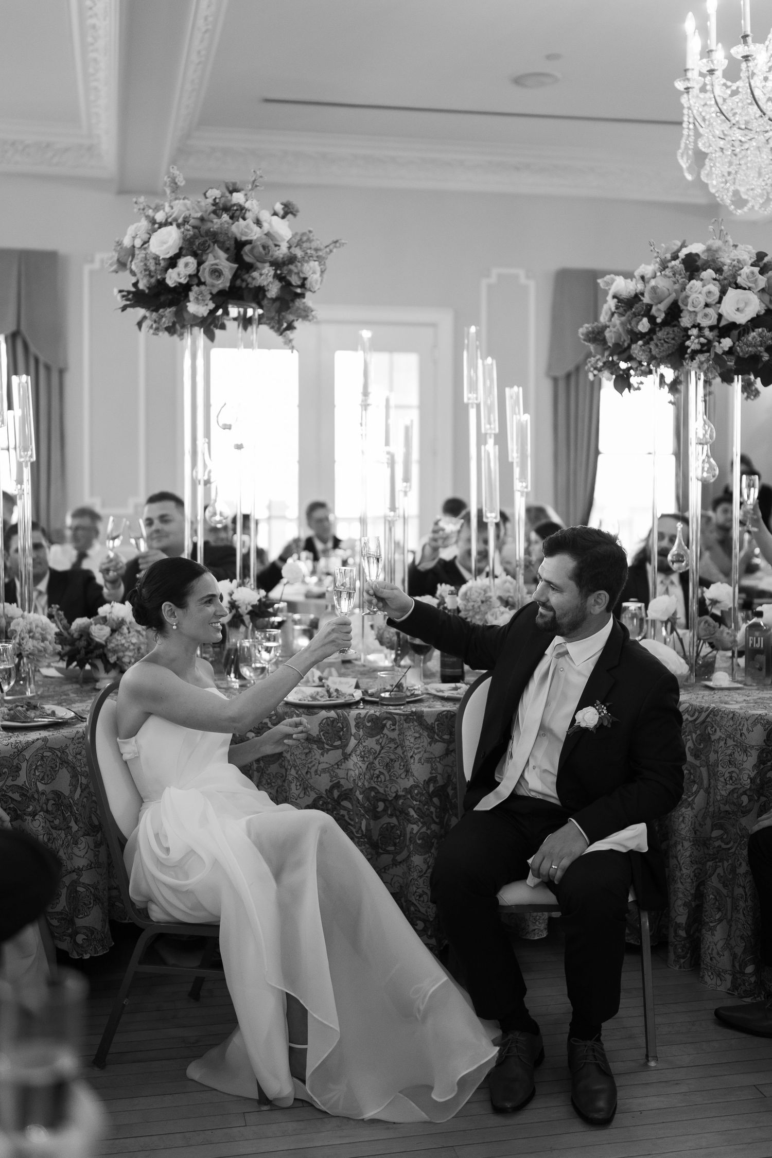 A wedding reception featuring a decorated dining table with tall floral centerpieces and crystal chandeliers in an elegant ballroom.
