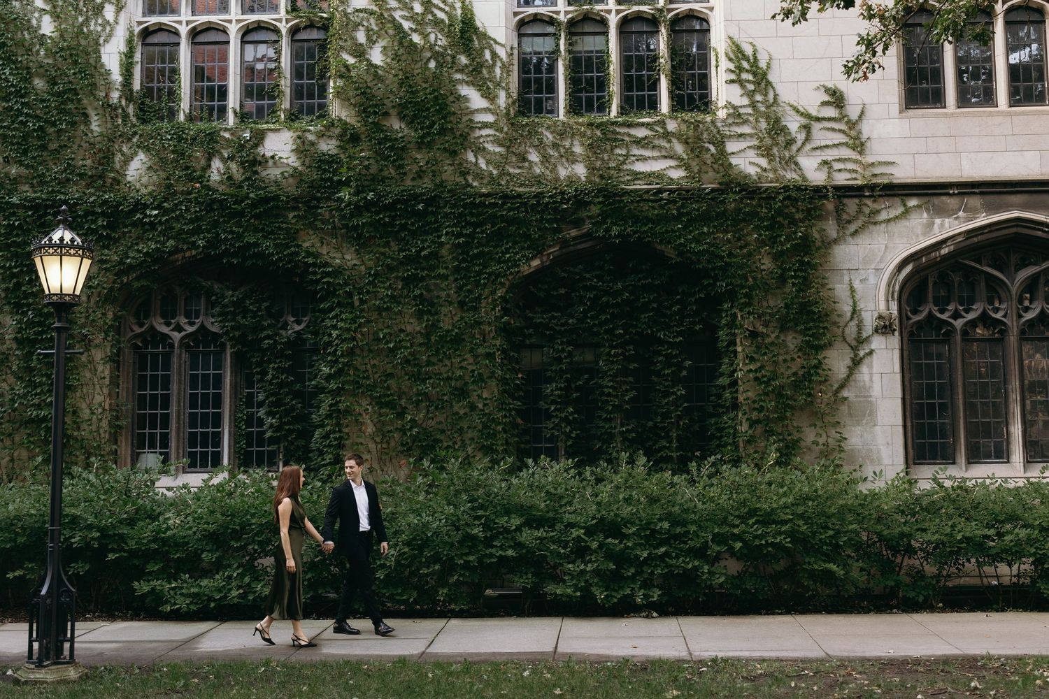 Two people walking along a path beside an ivy-covered collegiate Gothic building.