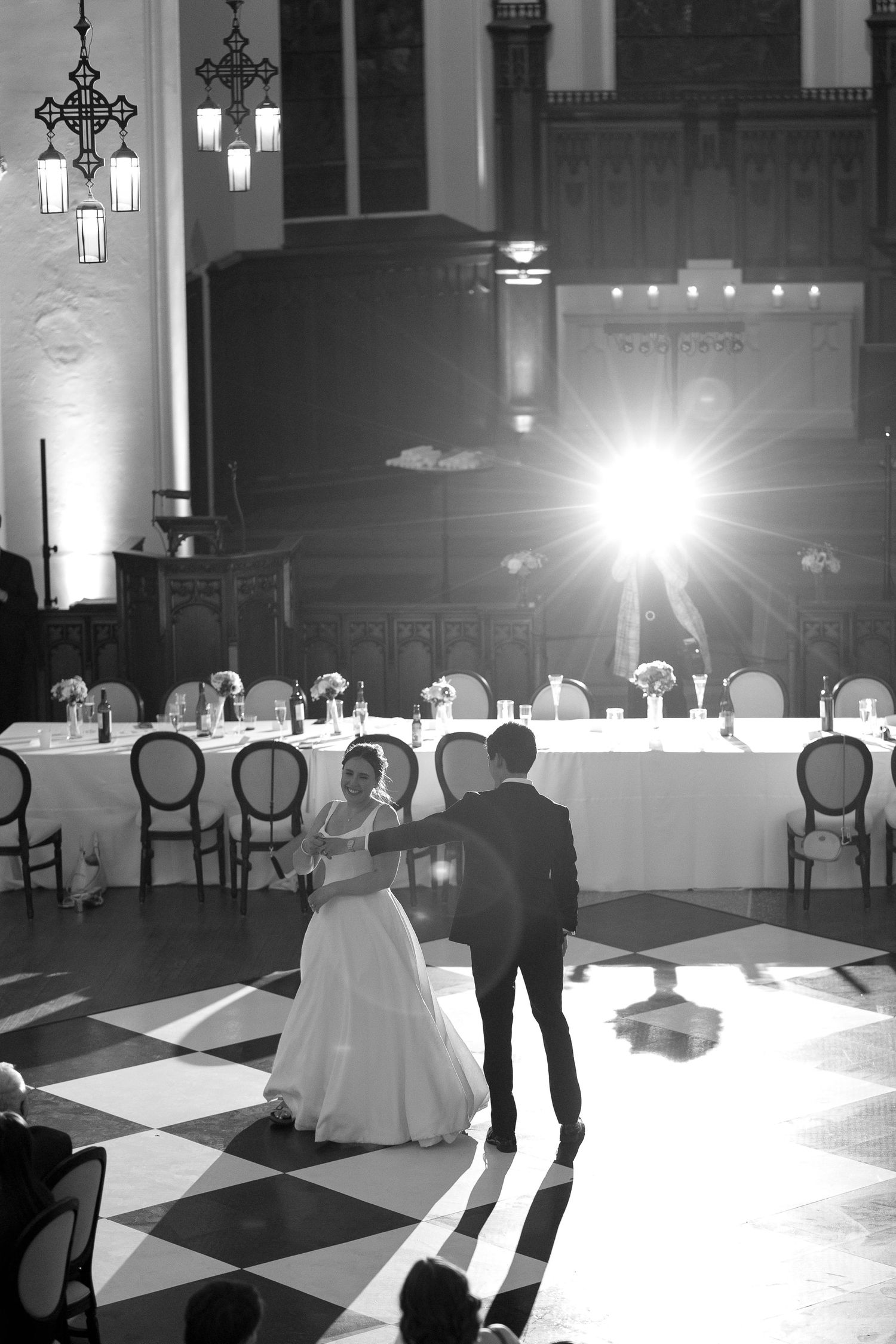 Couple dancing in spotlight on checkered ballroom floor during wedding reception.
