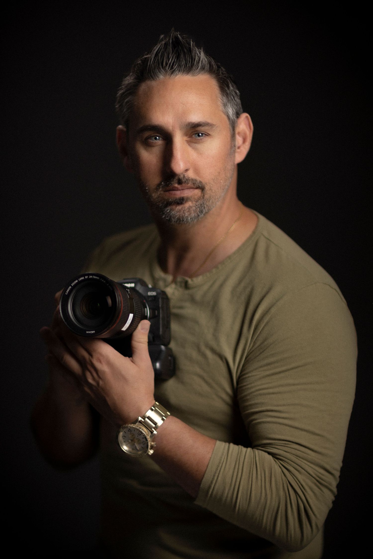 Professional photographer in olive green long sleeve shirt holds DSLR camera against dark background.