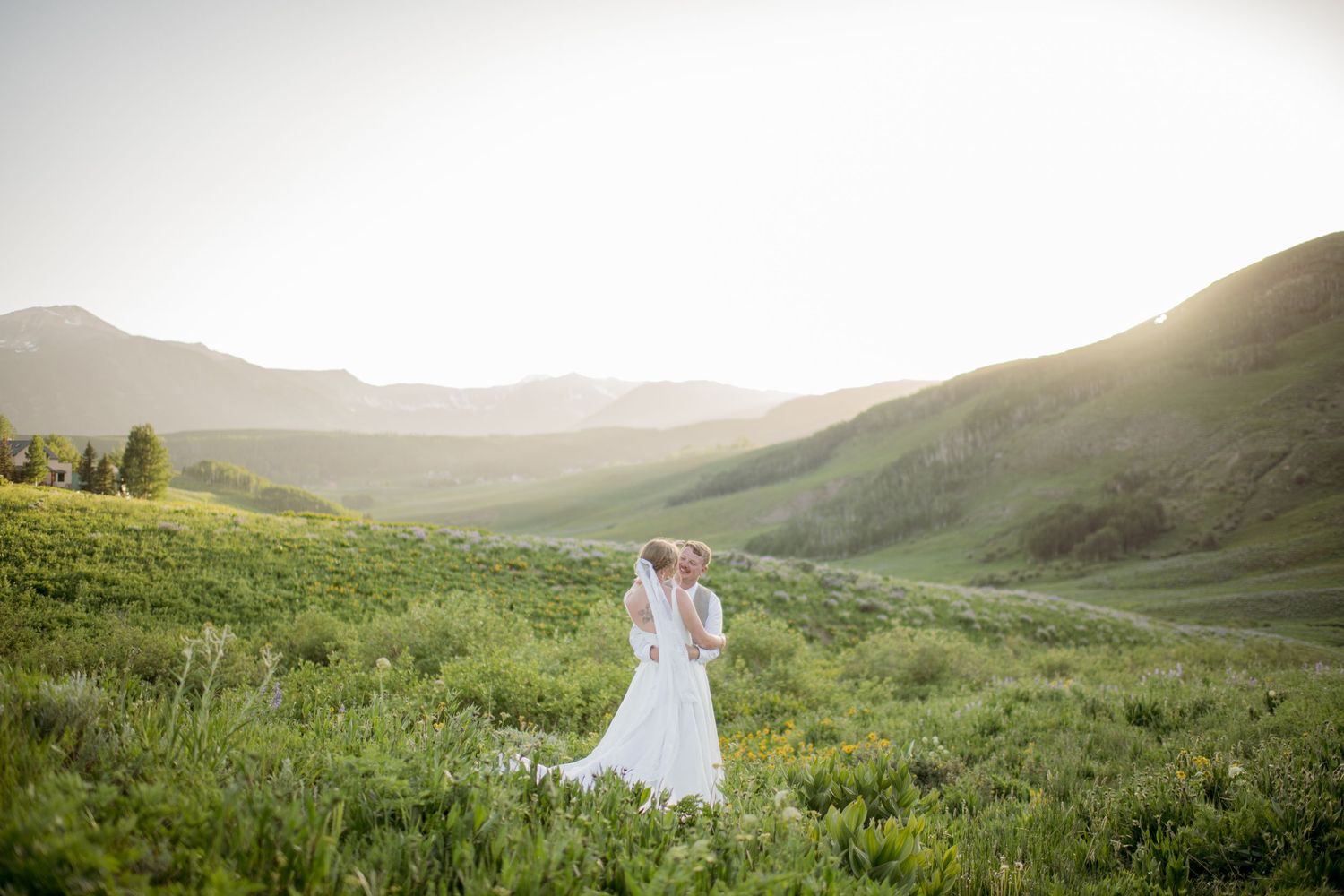 Couple embraces in meadow surrounded by rolling hills during golden hour sunset.