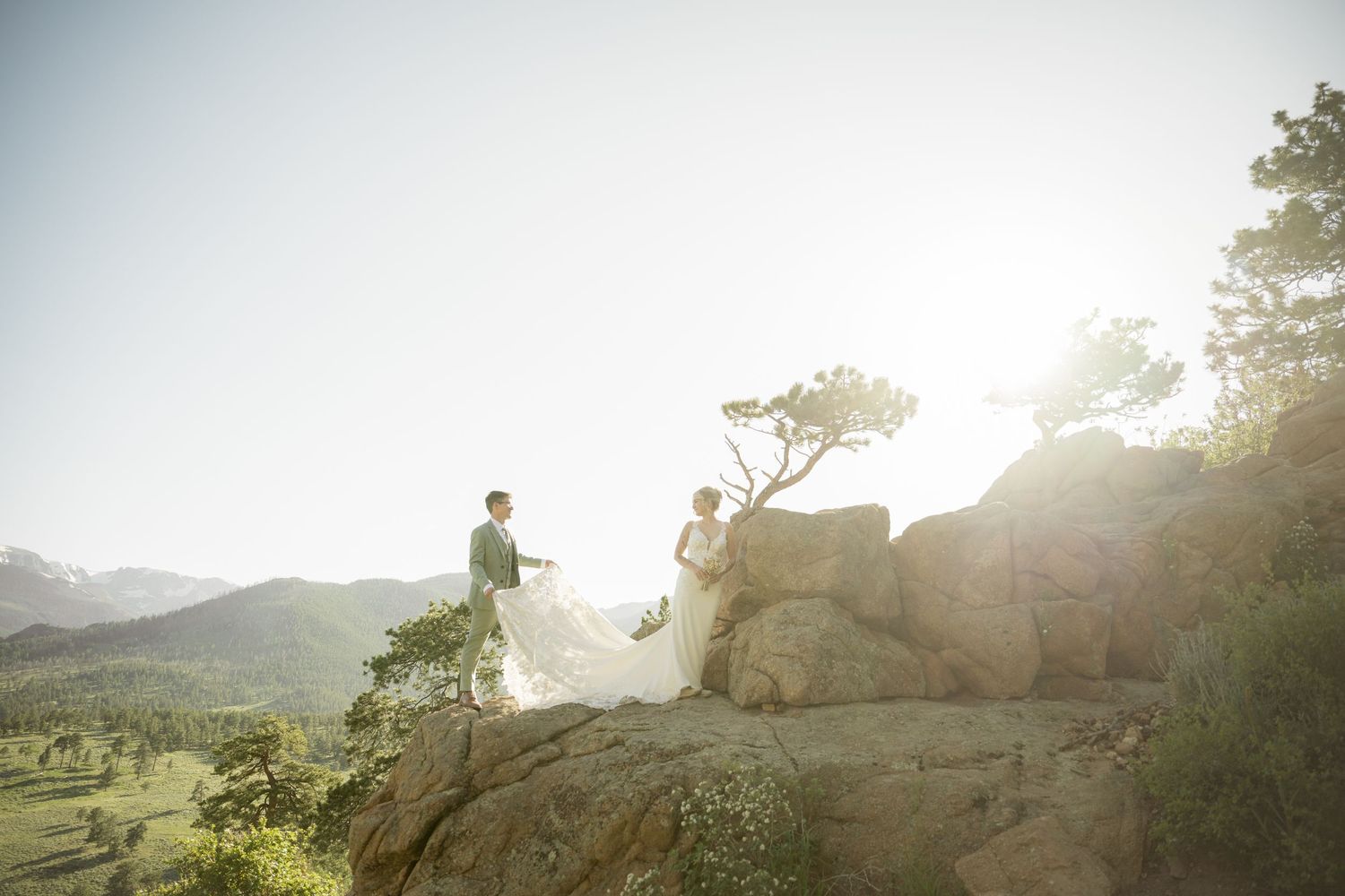 Ethereal wedding photoshoot silhouette against golden sunlight on mountain cliff with dramatic landscape view below.