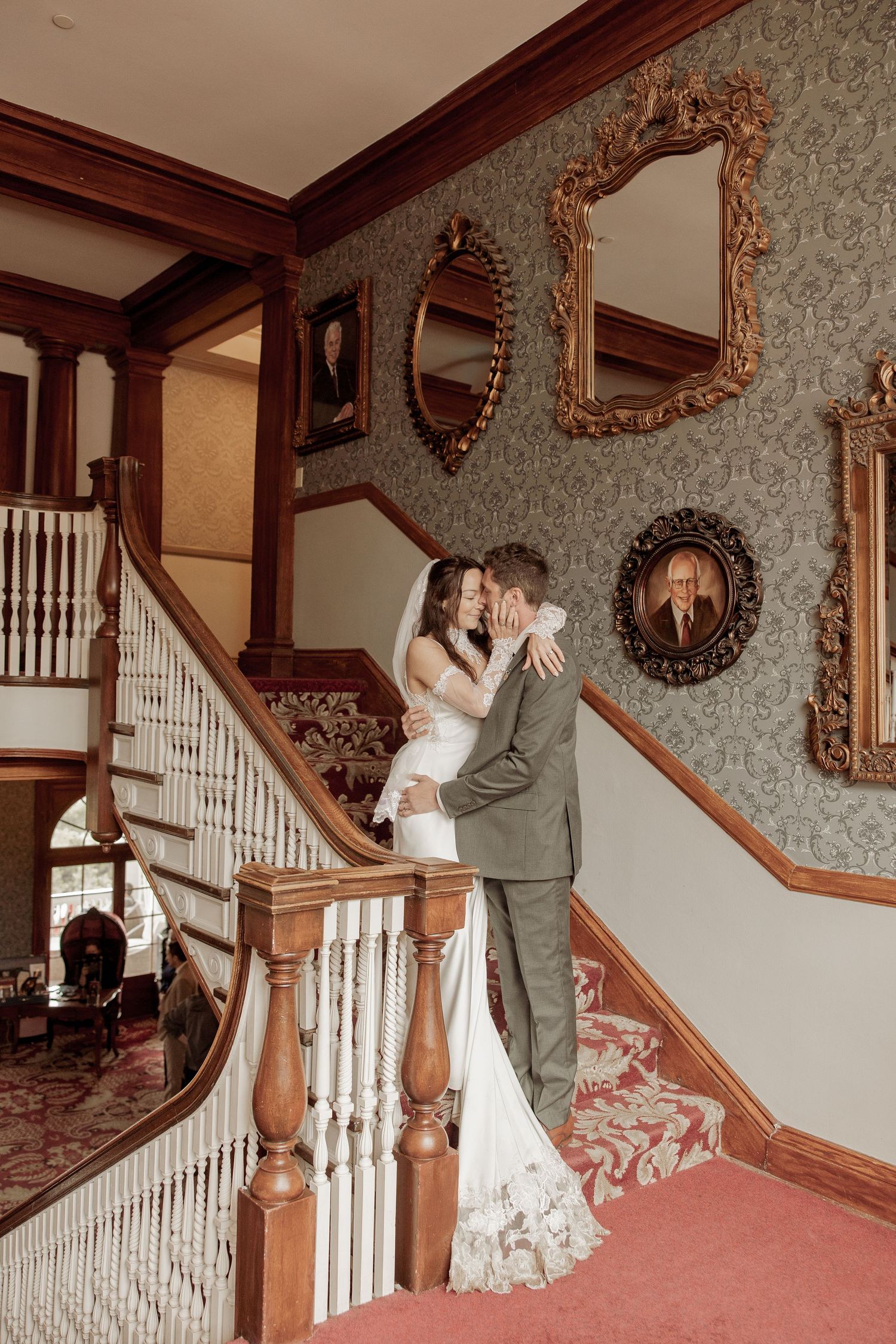 Romantic couple embraces on an elegant Victorian staircase with ornate wallpaper and vintage framed portraits in background.
