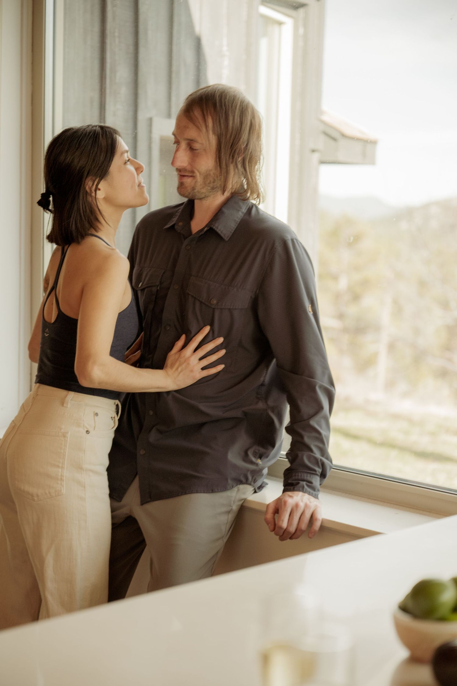 A romantic couple shares an intimate moment by a bright window in a modern kitchen interior.