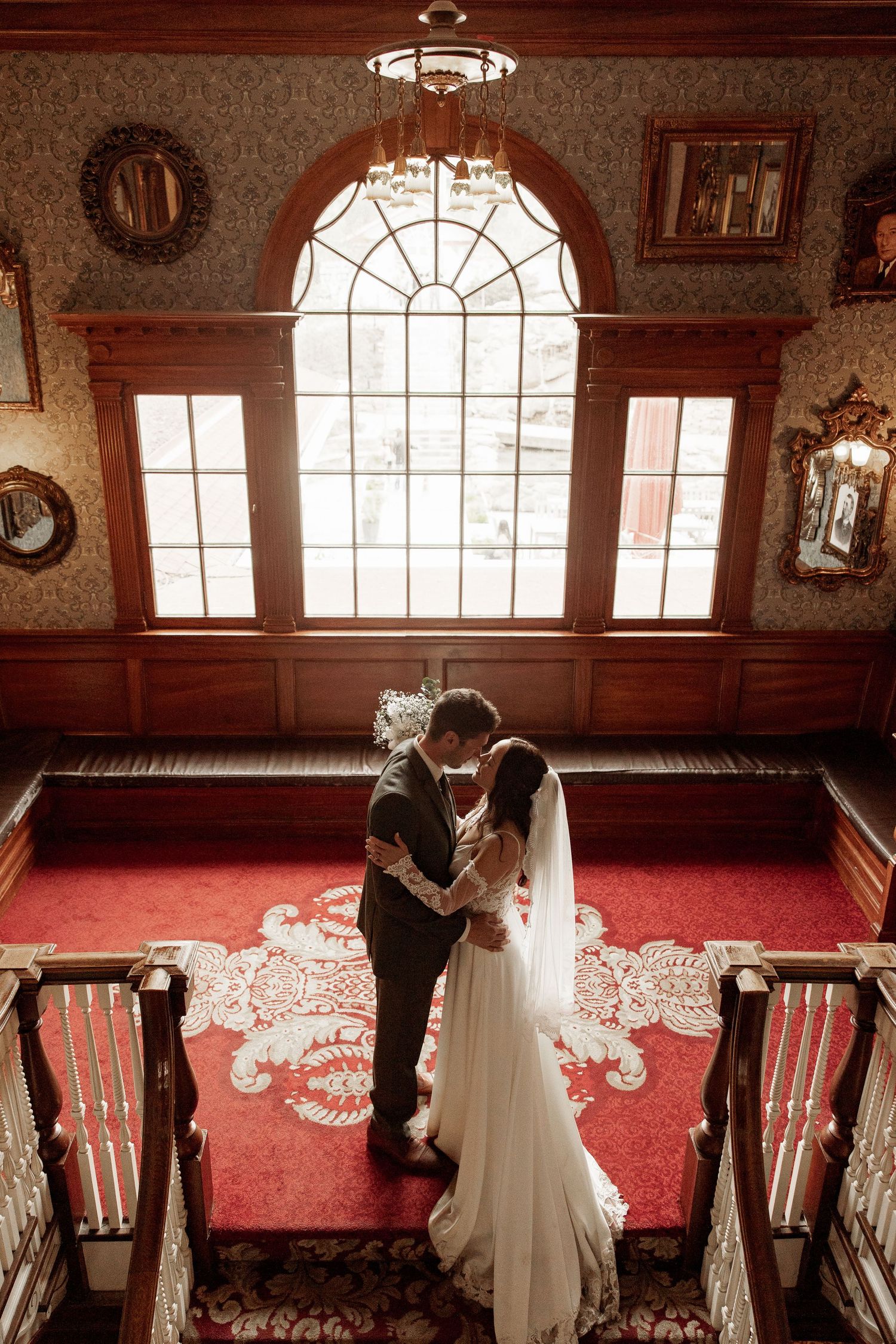 Romantic wedding scene in vintage mansion with arched window and red carpet on stairs, illuminated by natural light.