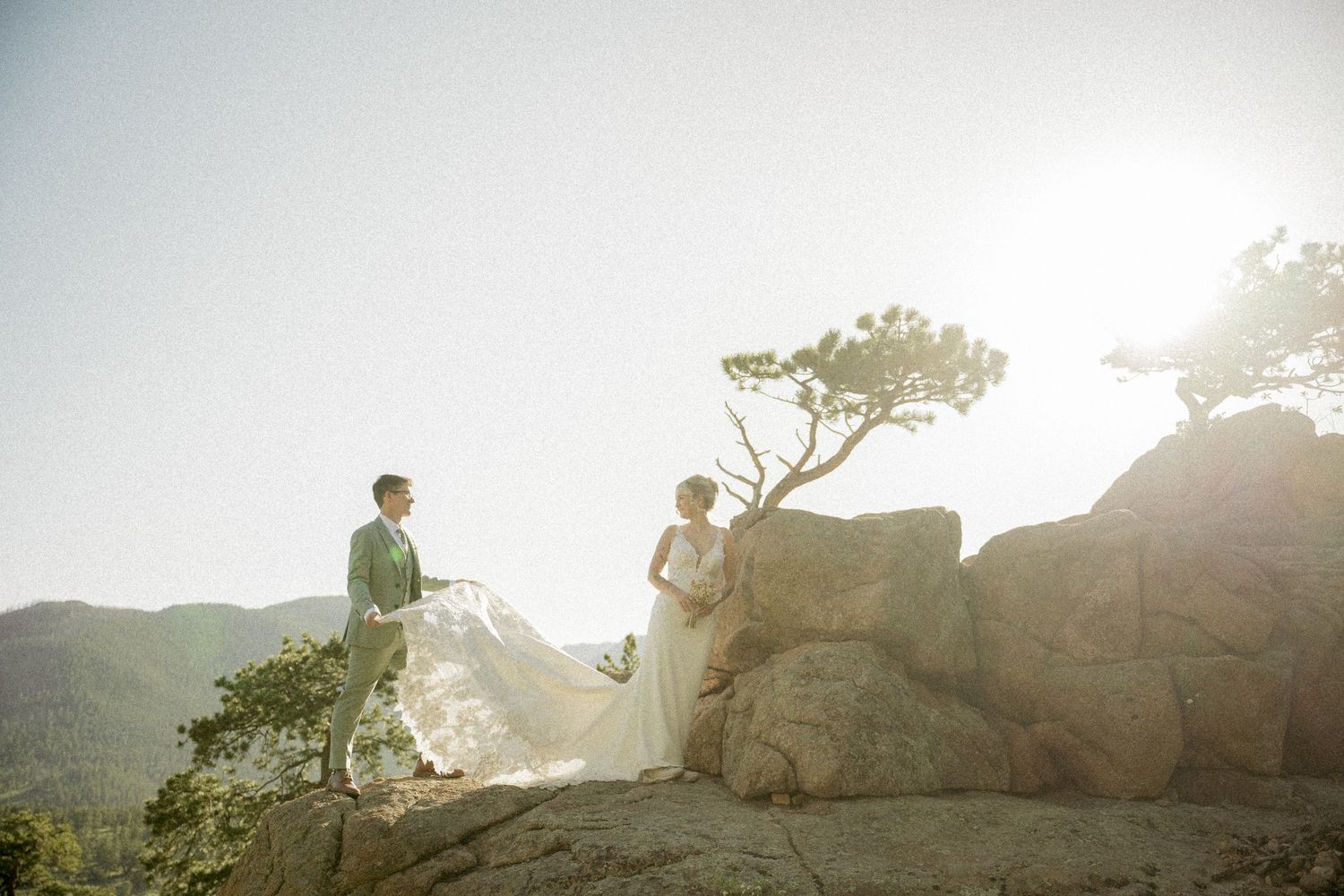 A couple stands atop a rocky mountain peak with windswept tree and dreamy backlit sunset atmosphere.