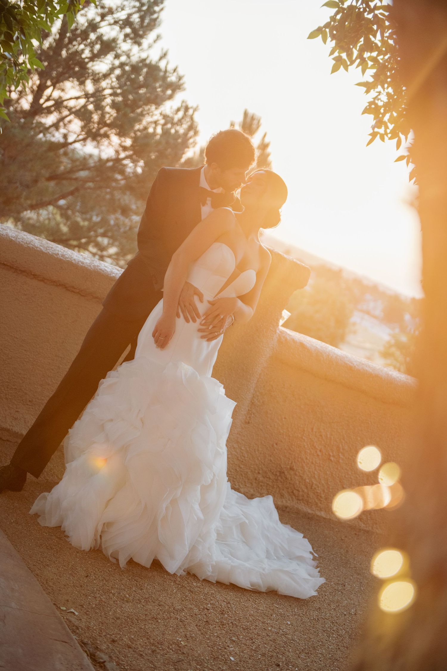 A romantic couple in wedding attire share an intimate embrace at sunset with beautiful golden light and lens flares.