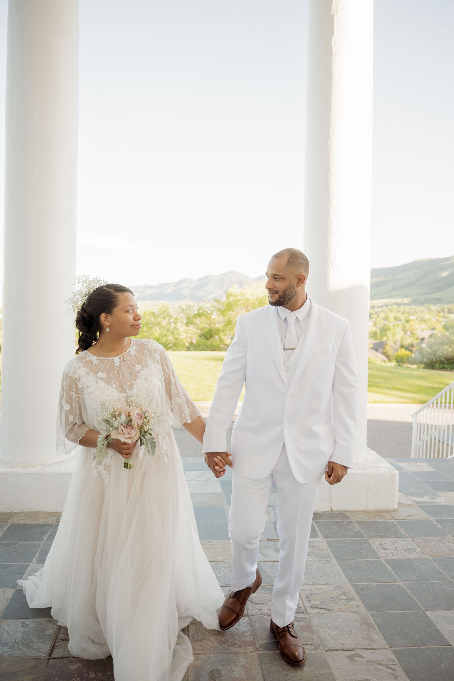 A romantic outdoor wedding portrait of a couple in white attire holding hands on a stone patio with mountain views.