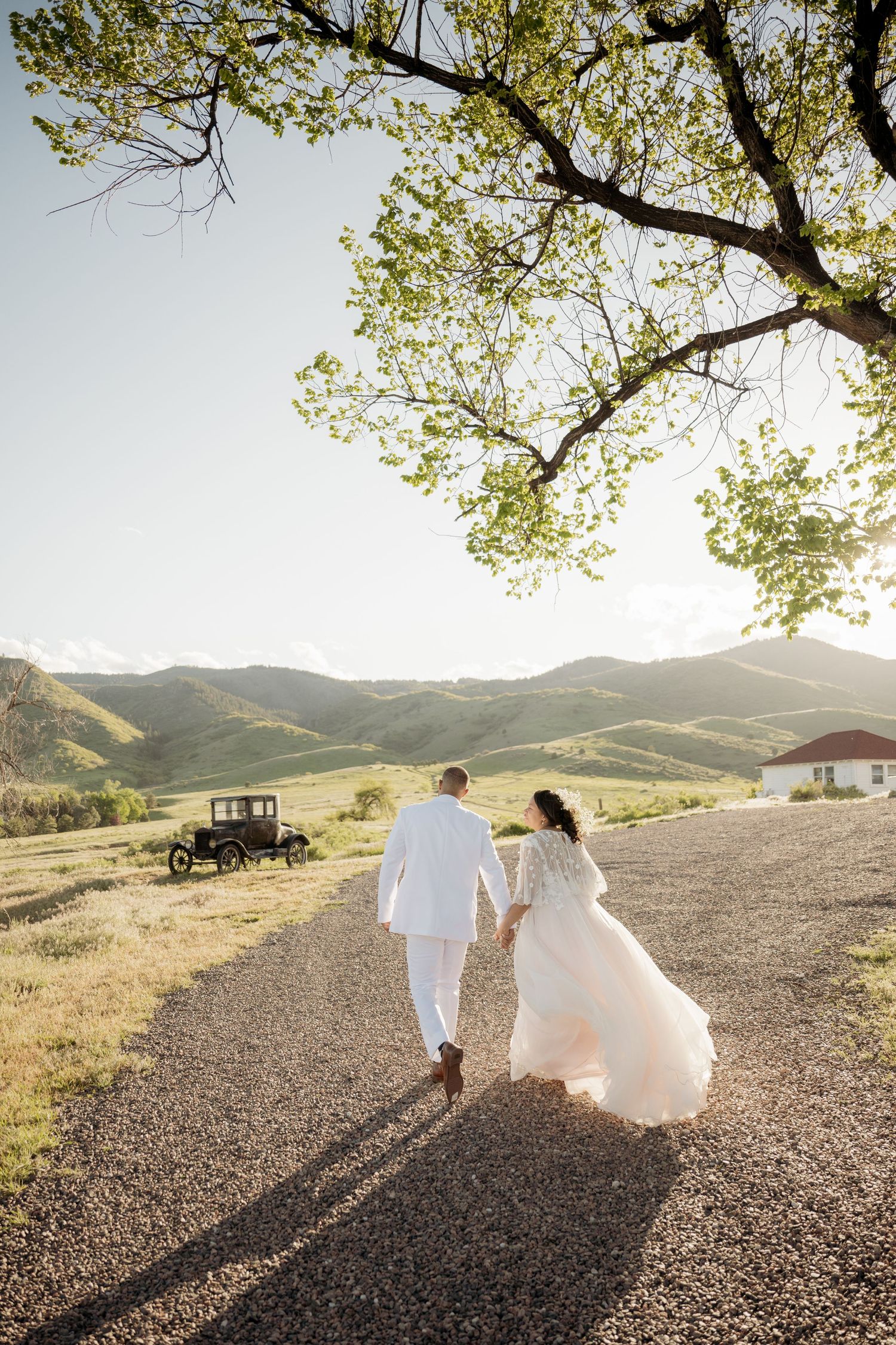 A couple walks hand in hand on a gravel path with rolling hills and a vintage car in the background during golden hour.