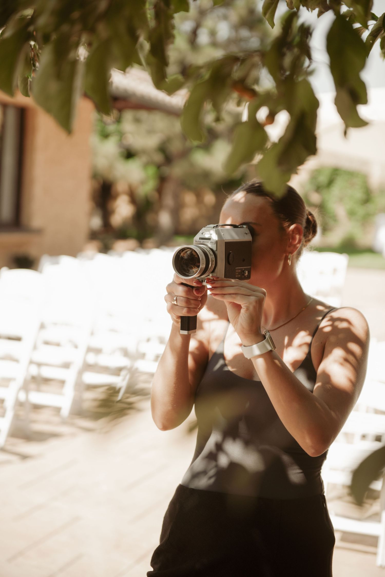 Professional photographer wearing a black dress takes pictures with vintage camera in an outdoor venue with white chairs.