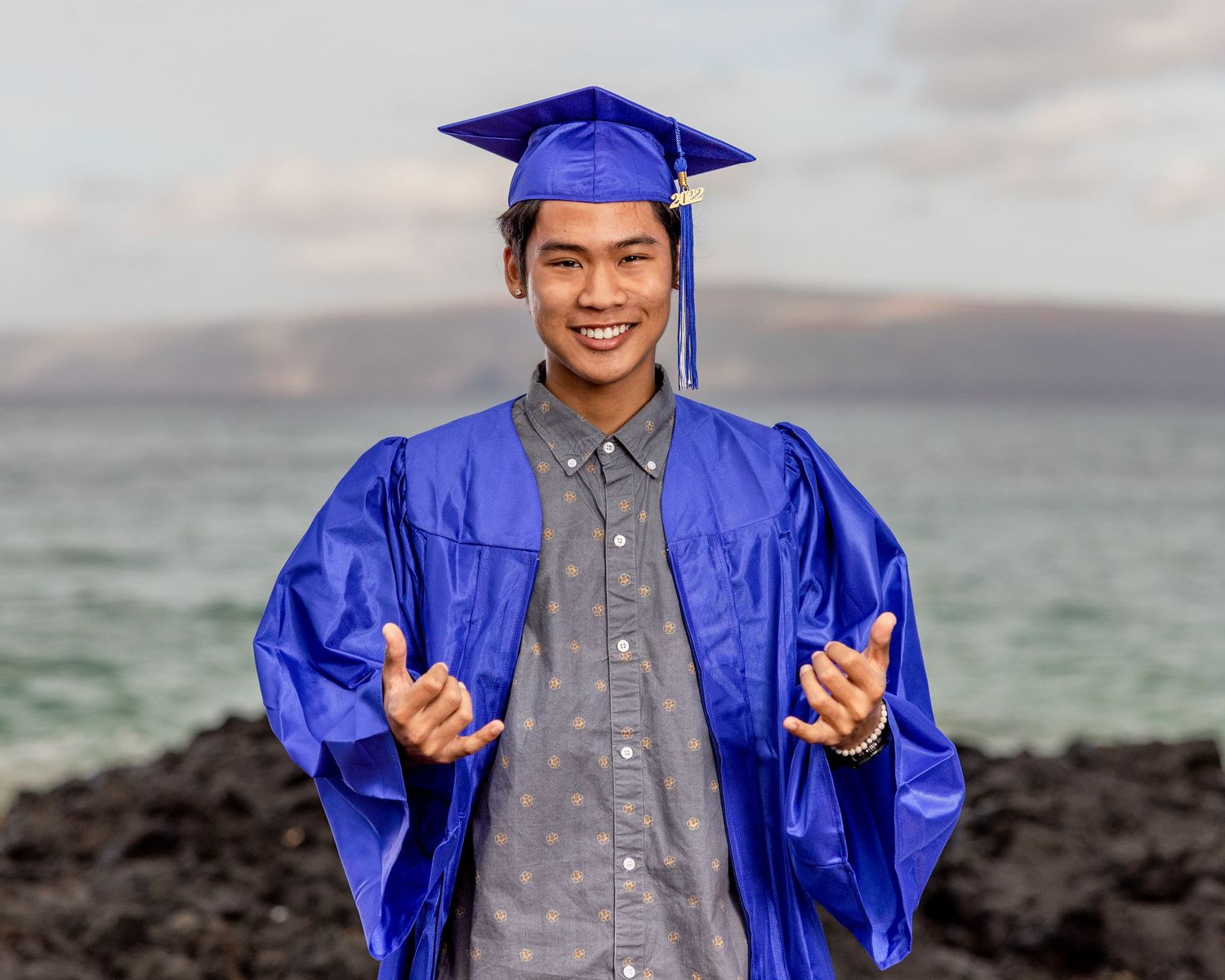 Graduate in blue cap and gown poses against scenic ocean background.