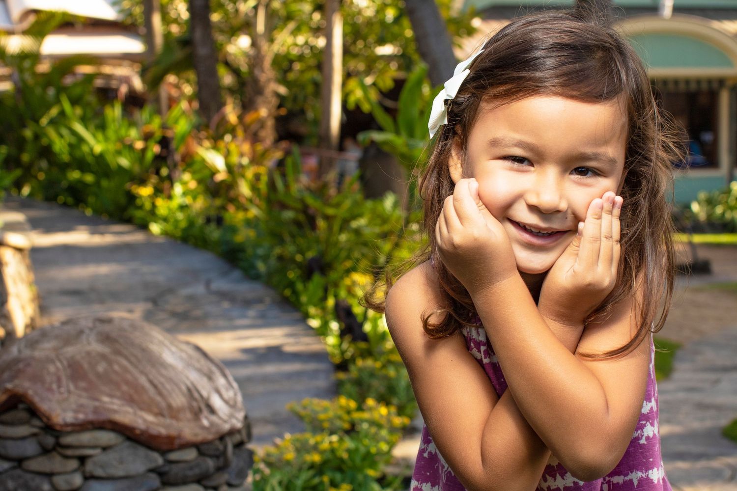 Child in purple dress smiling with hands on cheeks in a garden with greenery.
