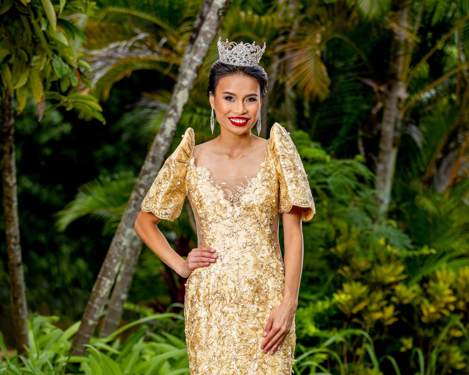 Pageant contestant in gold gown and crown poses elegantly in tropical garden setting.