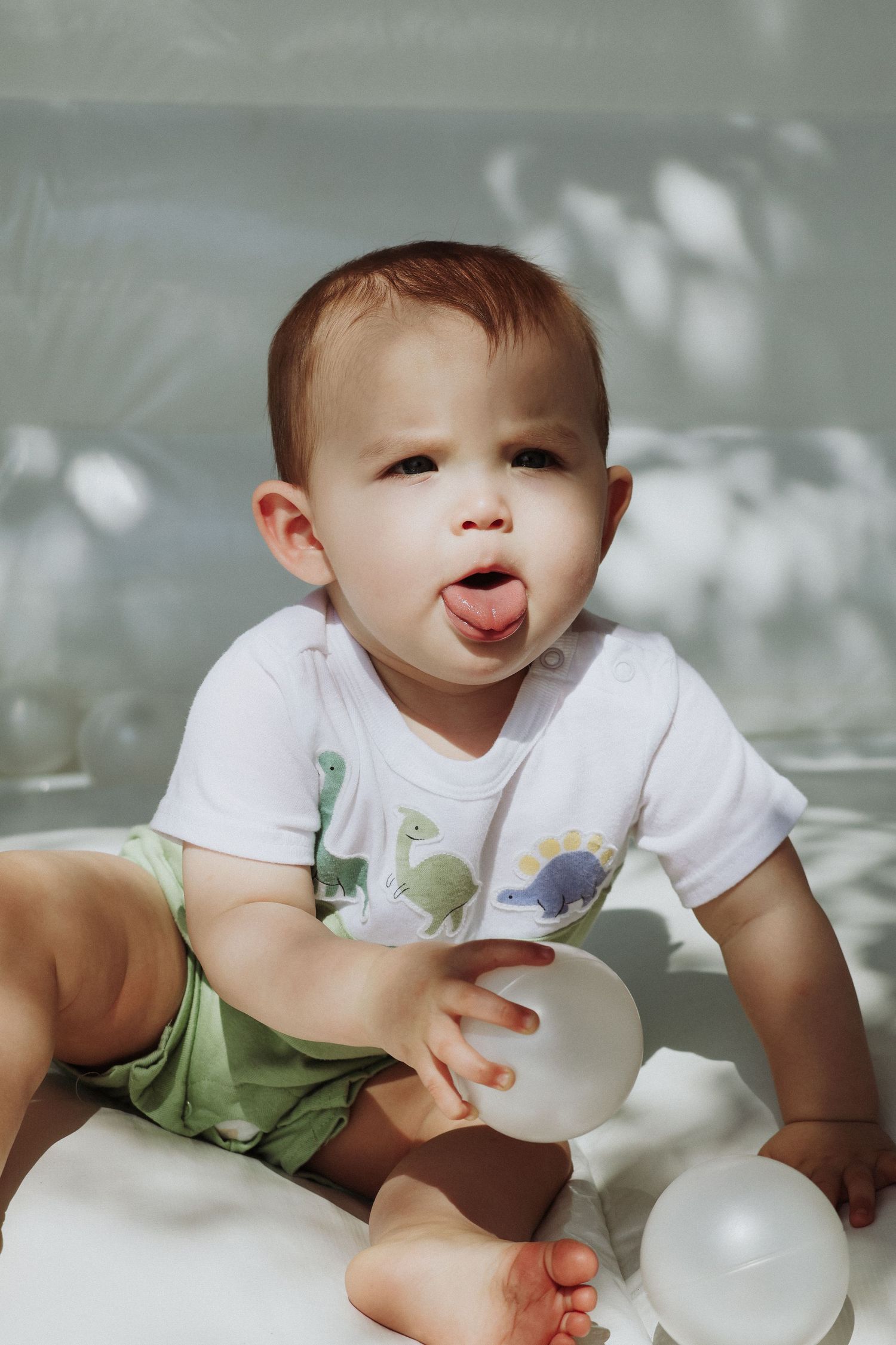 Playful baby sticking tongue out while sitting with white balloons in soft lighting.