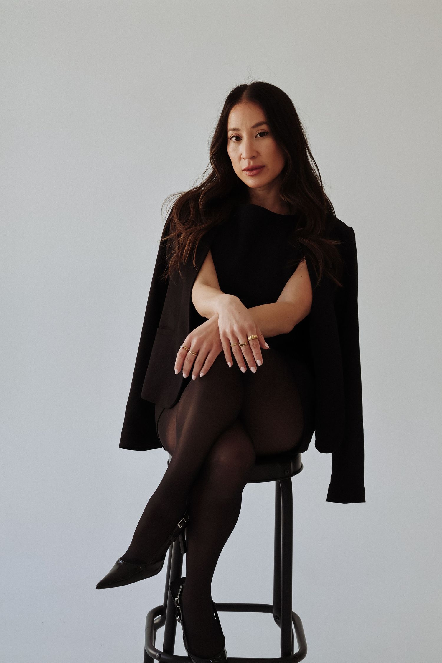 Model in black outfit posing on stool against white wall showing professional fashion photography style.
