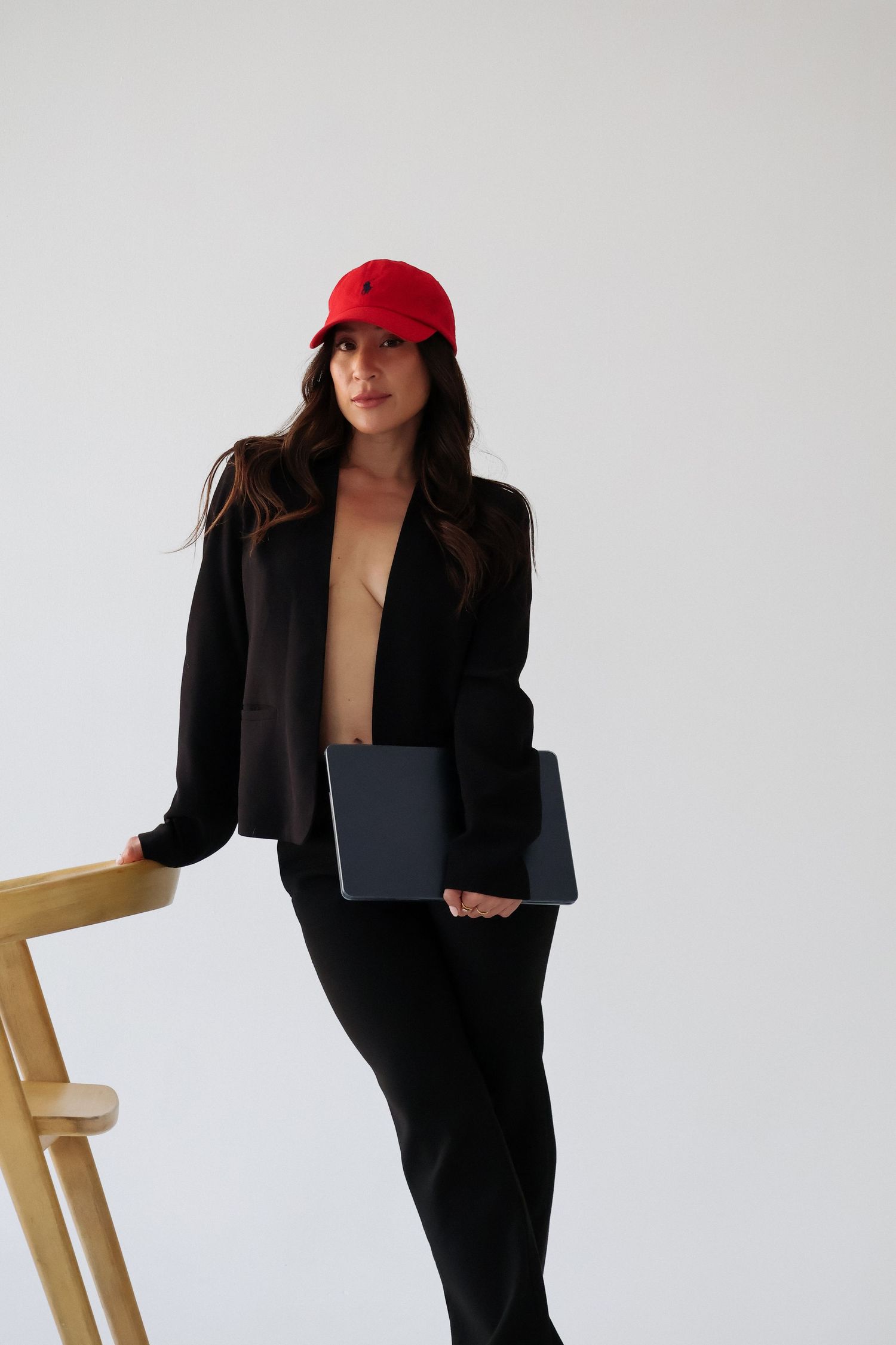 Professional woman in black suit and red cap poses with laptop against white wall.
