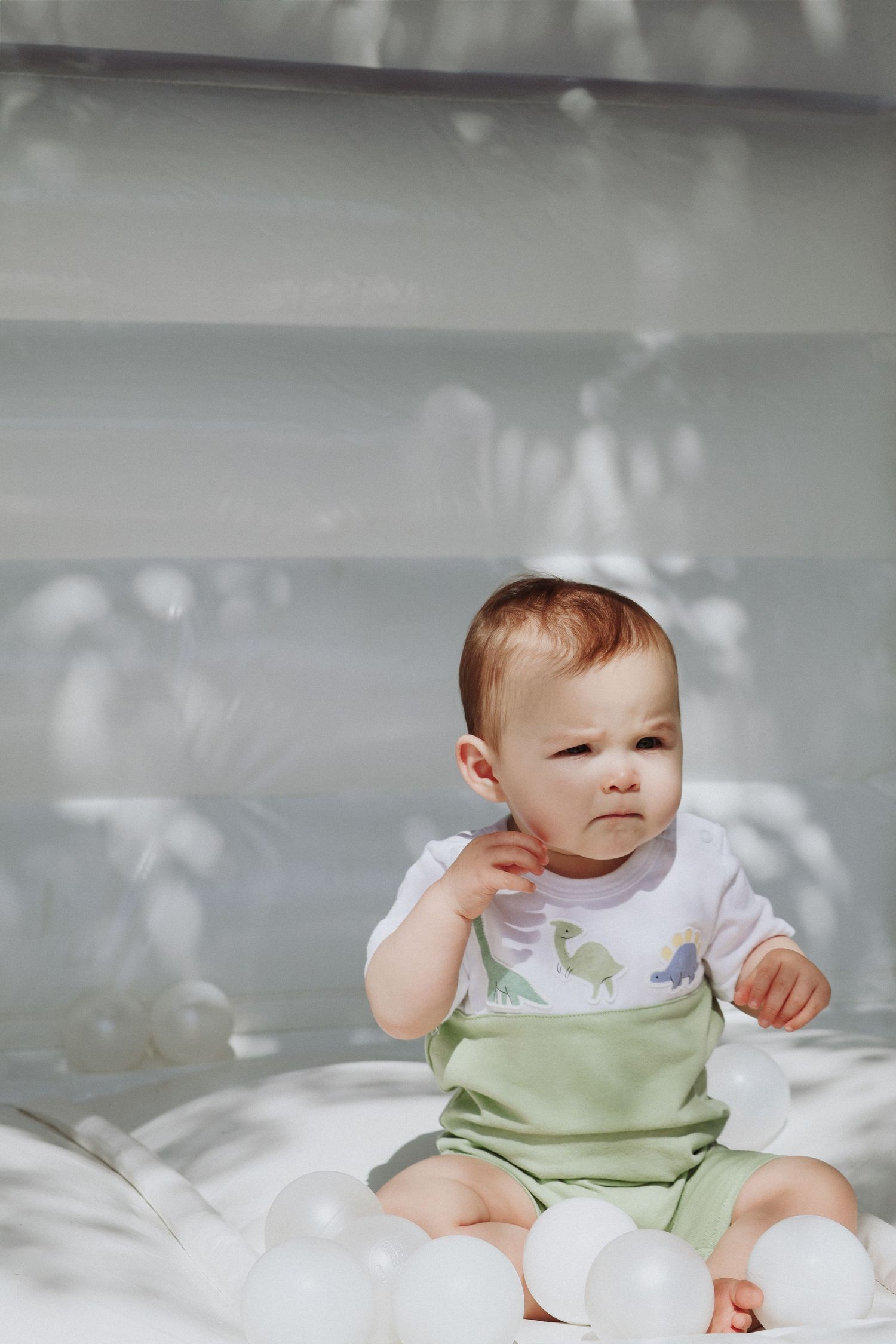 Baby in white and green outfit sitting on white bedding with dappled shadows.