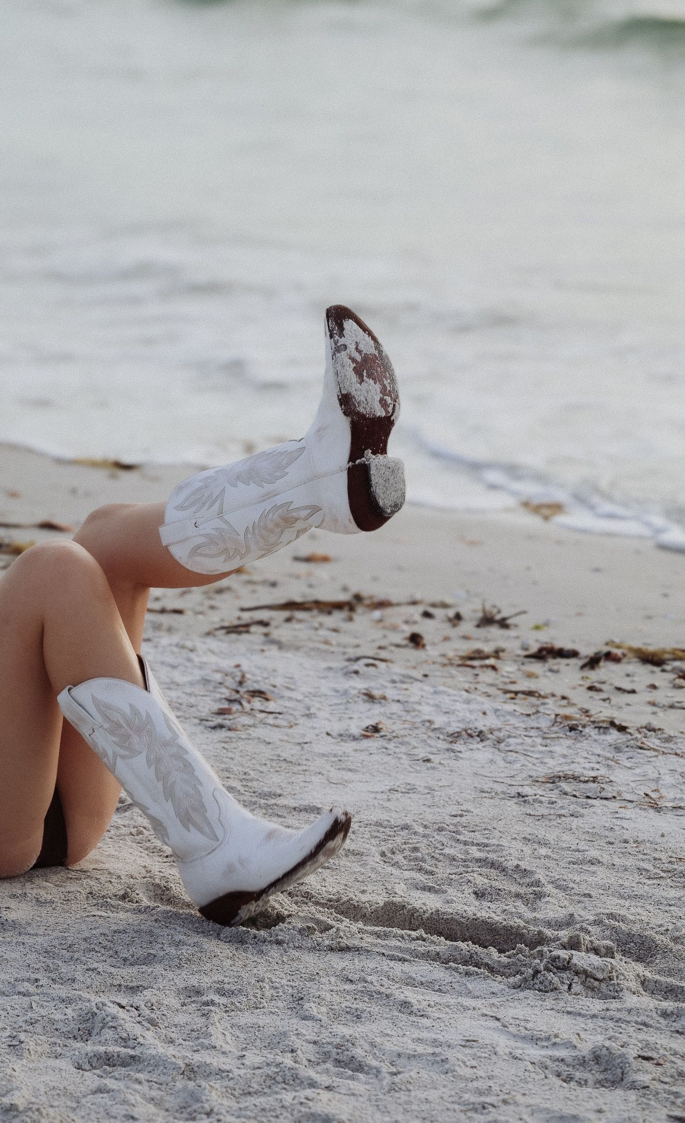 White cowboy boots resting on sandy beach with ocean waves in background.