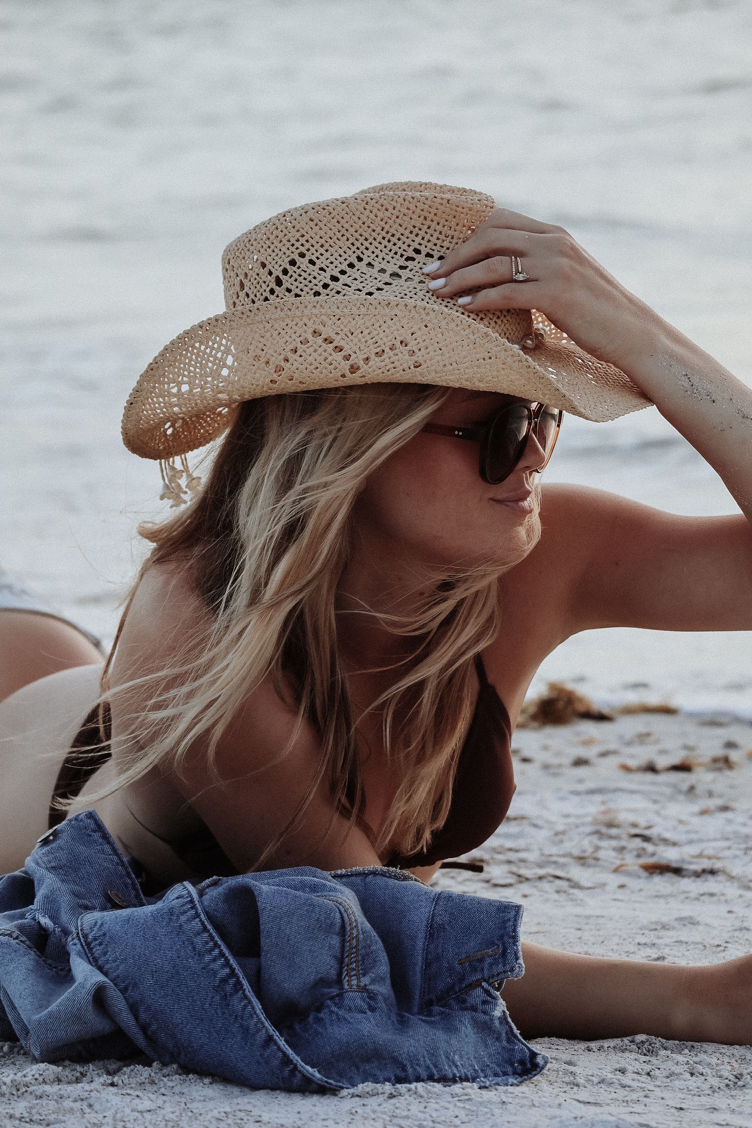 Woman lying on beach wearing straw hat and denim in artistic casual pose.