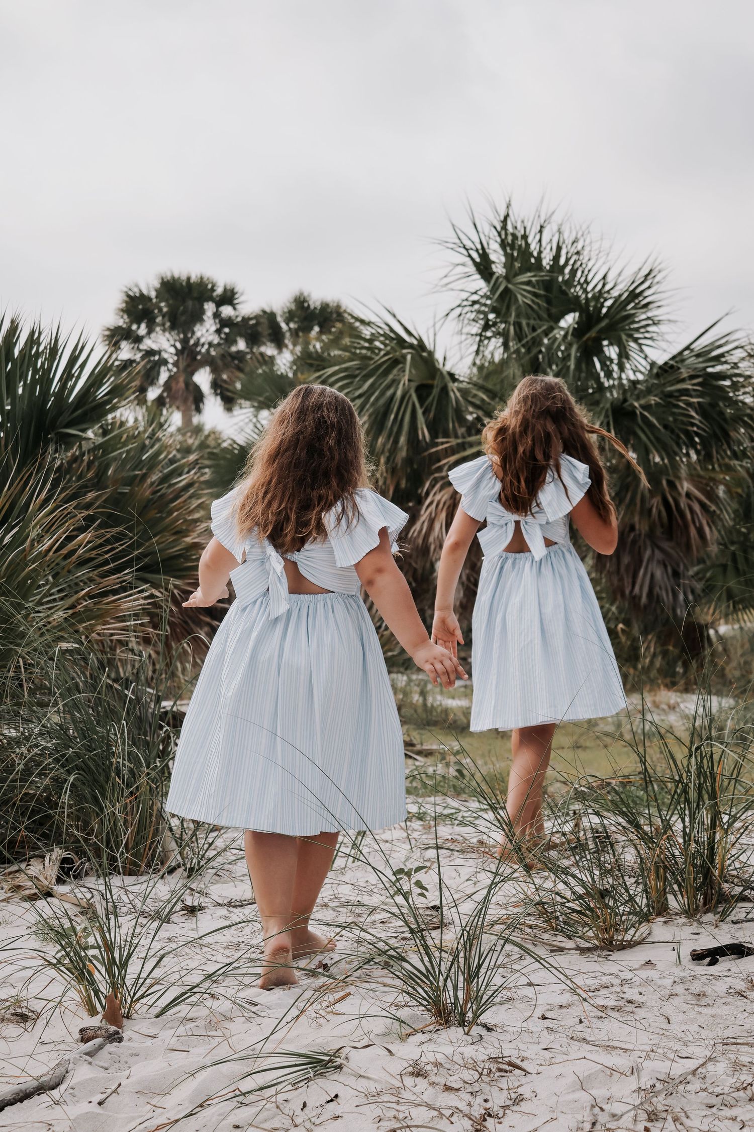 Two girls in matching white dresses walking hand in hand through beach dunes.