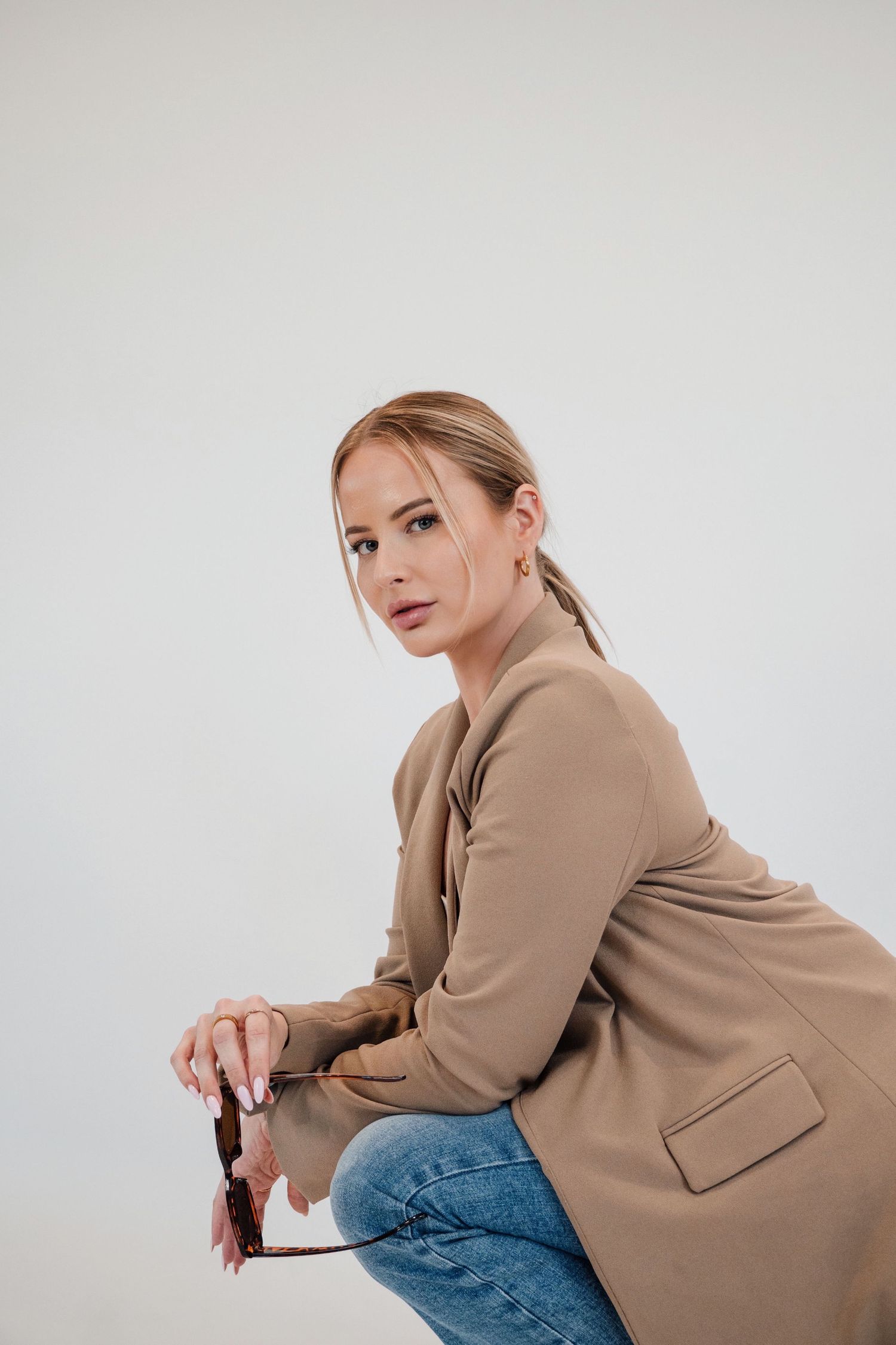 Fashion model in beige coat and jeans poses against white studio background.